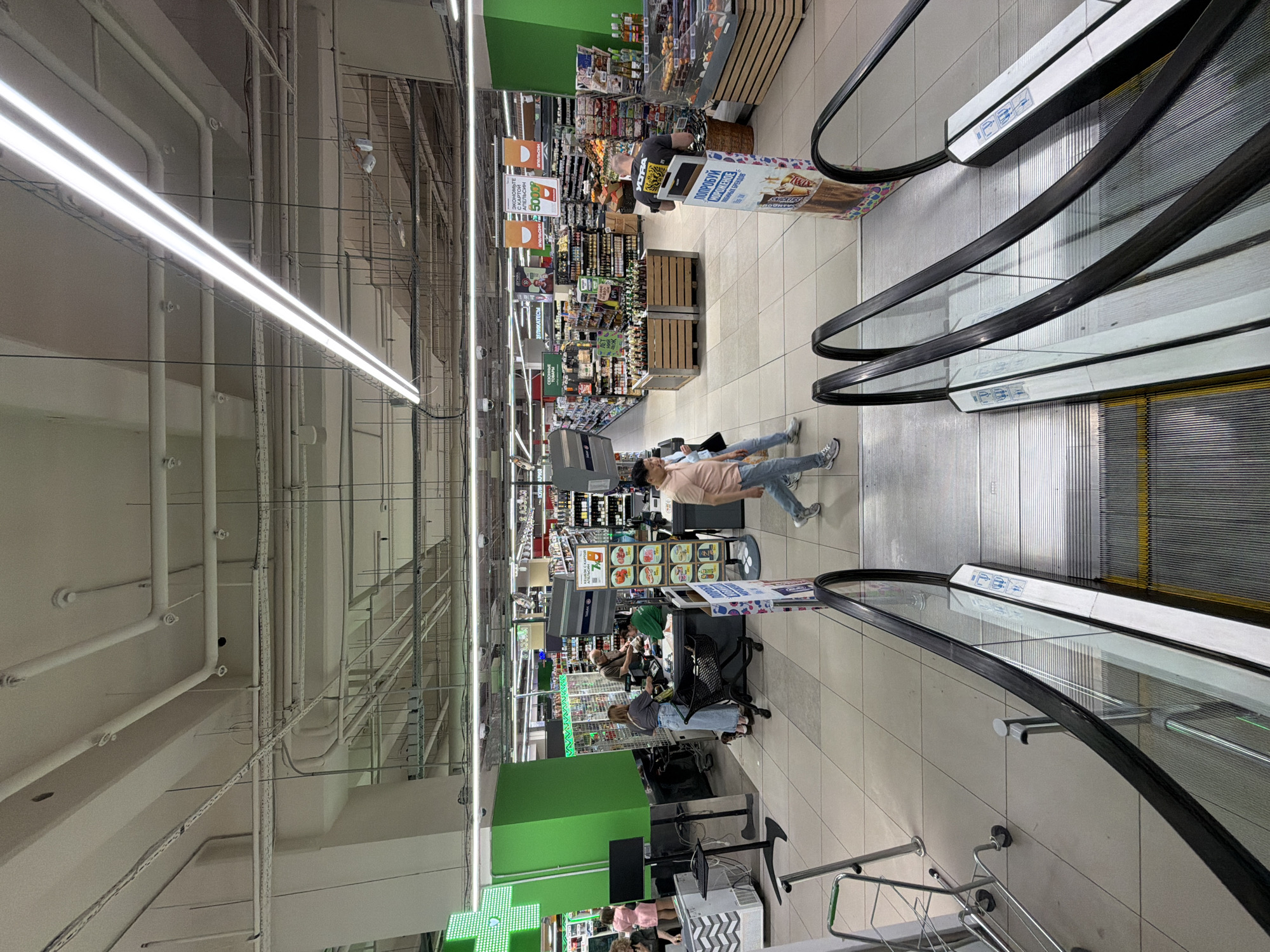 This image depicts the interior of a supermarket, viewed from the top of a downward escalator looking into the main shopping area. The scene is brightly illuminated by numerous overhead fluorescent strip lights.
The ceiling features exposed utility infrastructure, including a complex network of white pipes, electrical conduits, ventilation ducts, metal cable trays, and multiple security cameras. The floor is covered with large, light-colored square tiles.
In the foreground, two parallel escalators descend into the store. They have black handrails, clear glass side panels, and steps with a textured metal surface and yellow warning strips. Blue informational stickers are visible on the side panels. To the immediate left of the escalators, a silver shopping cart with green handles stands on the tiled floor.
Multiple individuals are present in the supermarket. In the mid-ground, centered slightly to the left, is a checkout area with two active registers. A woman in a light blue top and jeans, pushing a black wire shopping cart, is interacting with a male cashier wearing a brown shirt. Another female cashier in a green uniform stands behind an adjacent register. Further left, partially obscured by a large green pillar, a woman in a pink dress is visible. The green pillar features a prominent green cross symbol, suggesting a pharmacy or health-related section.
In the central aisle, a man in a dark grey t-shirt and jeans stands facing a tall promotional display stand for "МОРОЖЕНОЕ" (ice cream). Nearby, another man in a light-colored shirt and jeans walks past him. The aisle contains wooden crate displays filled with bottled beverages, likely beer or kvass, and various shelves stocked with groceries.
Overhead, several large orange banners are suspended, one clearly displaying "ЭКОНОМЬТЕ С КАРТОЙ АПЕЛЬСИН" (Save with Orange card) and another showing "500P" (500 Rubles). Additional signage in Russian is visible throughout the store, including "ДЕЛ ИКАТЕСЫ" (Delicacies) and "СЕЗОННЫЕ ТОВАРЫ" (Seasonal products). A refrigerated display case with pastries or desserts is located on the right side of the main shopping area, alongside sections for fruits and vegetables. In the distant background, faint Russian text including "КАЗАНЬ" (Kazan) is discernible on a banner.
The store's branding and language identify it as a Russian supermarket, consistent with chains found in Moscow, Russia.
