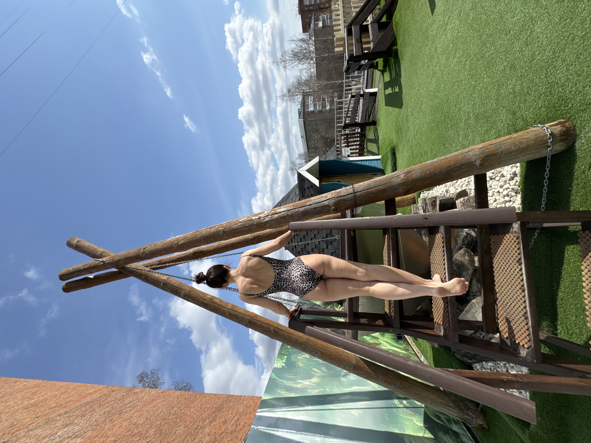 A woman in a leopard-print swimsuit is lying on a wooden structure in Votkinsk, Russia. The structure appears to be a decorative installation, possibly part of a playground or recreational area. It consists of a wooden frame supported by logs, with a metal staircase-like element. She is resting on her back, supported by the structure. The background features a green artificial turf surface, a small blue and white structure, and a building in the distance. The sky is blue with scattered clouds. The image is taken during daylight, with clear visibility.
