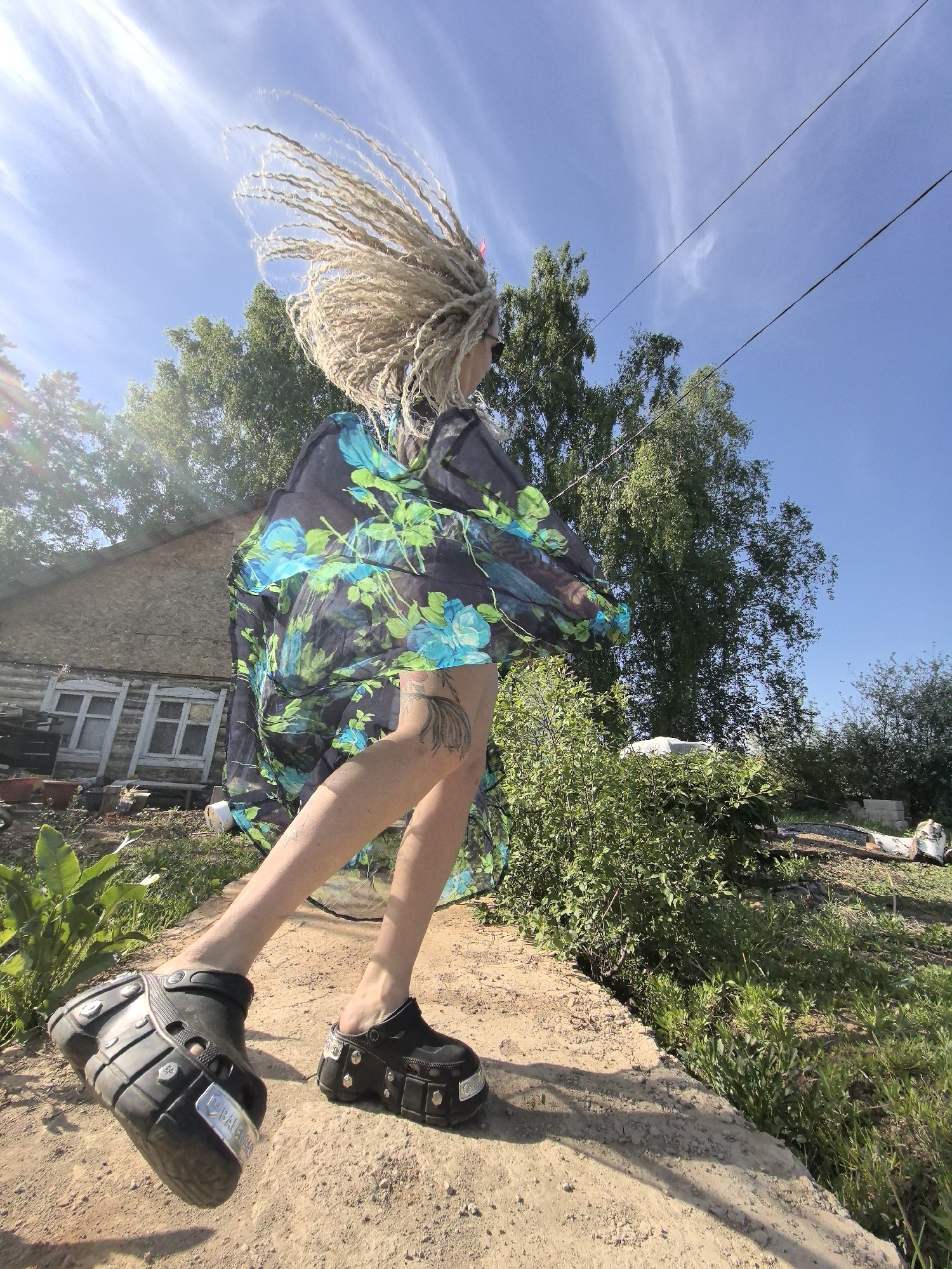 A woman with blonde dreadlocks and wearing a black and blue floral dress is walking on a dirt path in Noviy, Russia. She is wearing black platform Crocs. The sky is blue with some clouds. Behind her is a wooden building and greenery. Overhead power lines are visible. The scene is outdoors and sunlit.