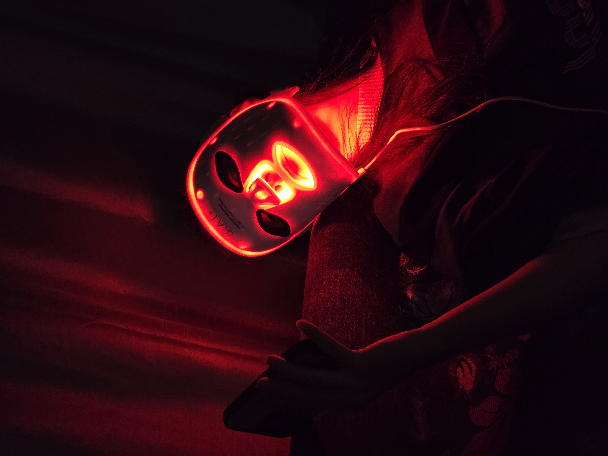 The image captures a person using a red LED light therapy face mask in a dimly lit, dark setting, likely indoors at night. The dominant light source is the intense red glow emanating from the mask, which illuminates the person's face, hair, and parts of their body and immediate surroundings.
The subject is seen from a slightly elevated, side-angle perspective, with their head tilted. Their long, dark hair is fanned out and highlighted by the red light. The white LED mask, designed with cutouts for eyes, nose, and mouth, covers the person's face. The red light within the mask creates a bright, almost fiery effect around the facial features.
On the lower left side of the mask, several lines of text are visible: "8 A+", followed by "LED Light Therapy Face Mask" and "Beauty Instrument". Below these, smaller, less legible text also appears.
The person appears to be reclining or lying down, with one hand visible in the lower right foreground. This hand is holding a dark, rectangular object, likely a smartphone, suggesting a moment of relaxation or multitasking during the treatment. The red light casts soft reflections on the person's skin and clothing, which appears to be dark fabric. The overall mood is futuristic and somewhat mysterious due to the intense monochrome red lighting against the dark background.