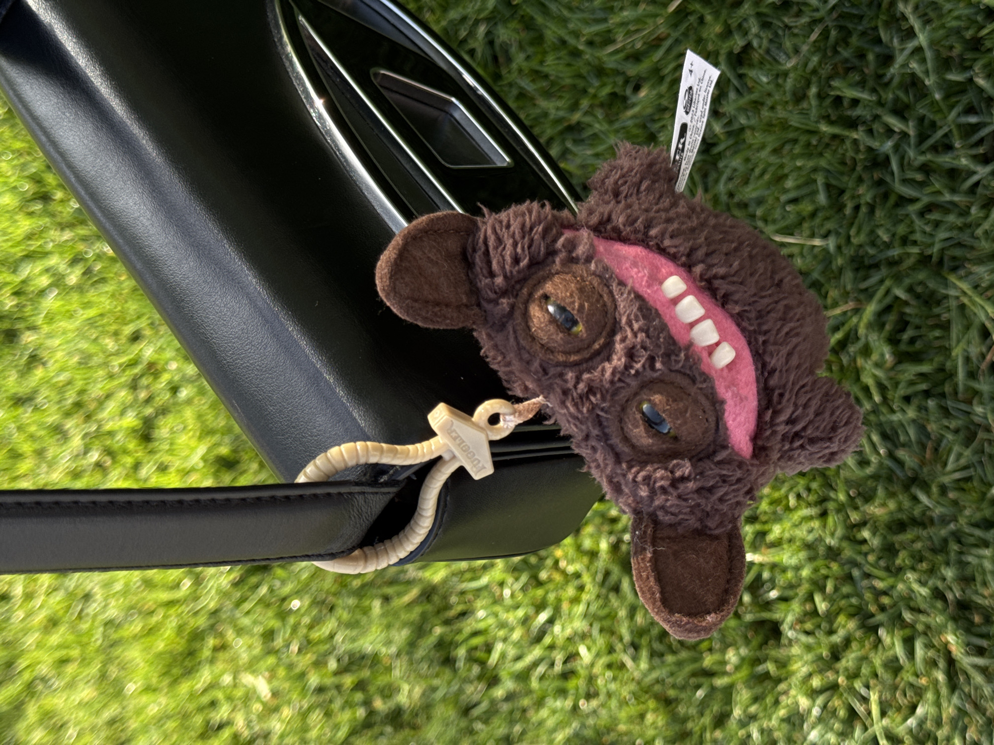 A brown, plush, monster-like toy with large eyes, a pink mouth, and white teeth is attached to a black leather purse with a white, plastic "YUGGSTER" tag. The purse is resting on green grass. The location is Belgrade, Serbia.