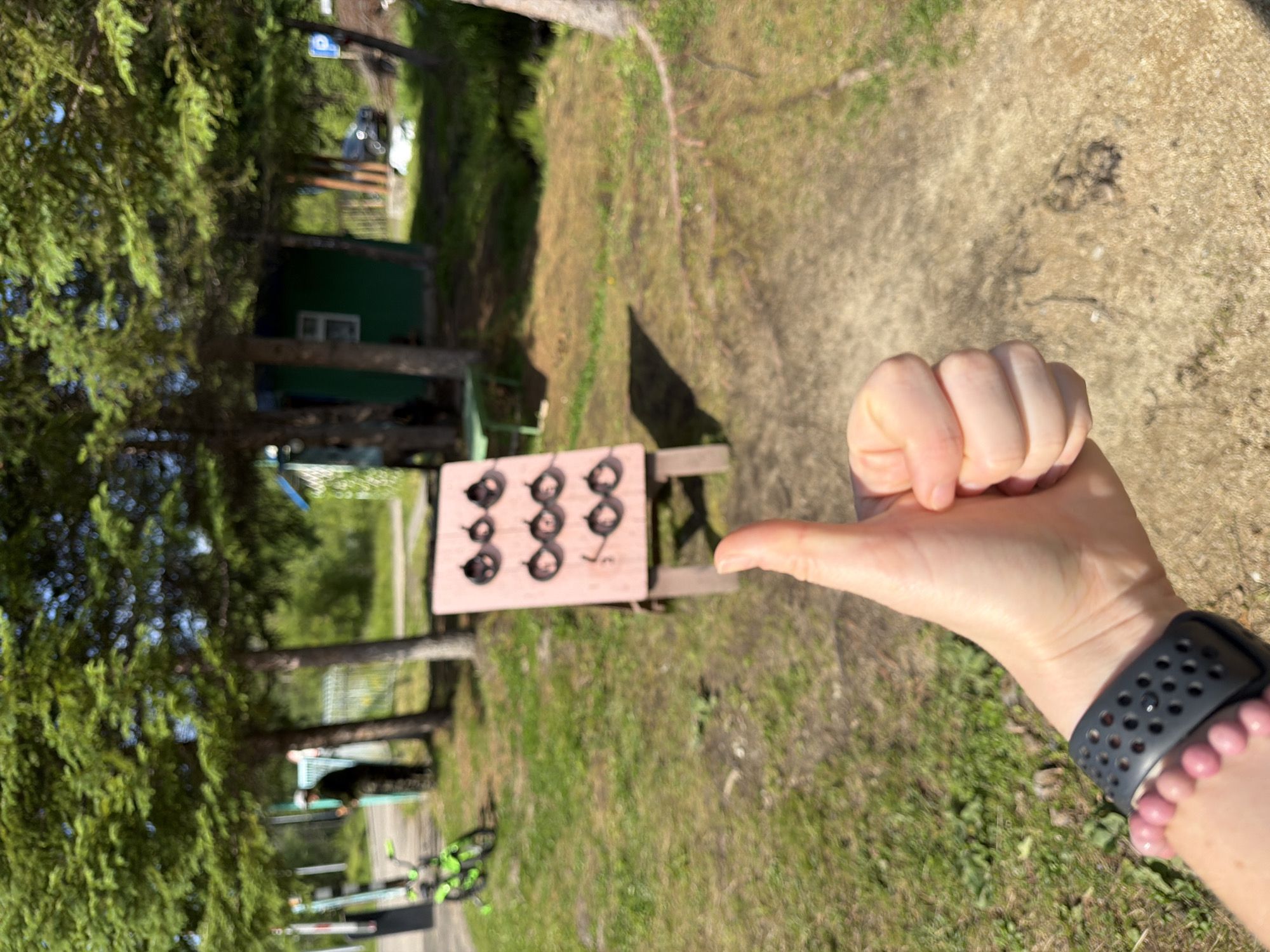 A person's hand gives a thumbs-up in the foreground. Behind it, a wooden board with rings on pegs is set up for a game. In the background, a small green building, trees, and a bicycle are visible. The scene appears to be outdoors in a rural or park-like setting, possibly in or near the city of Kul'dur, Russia.