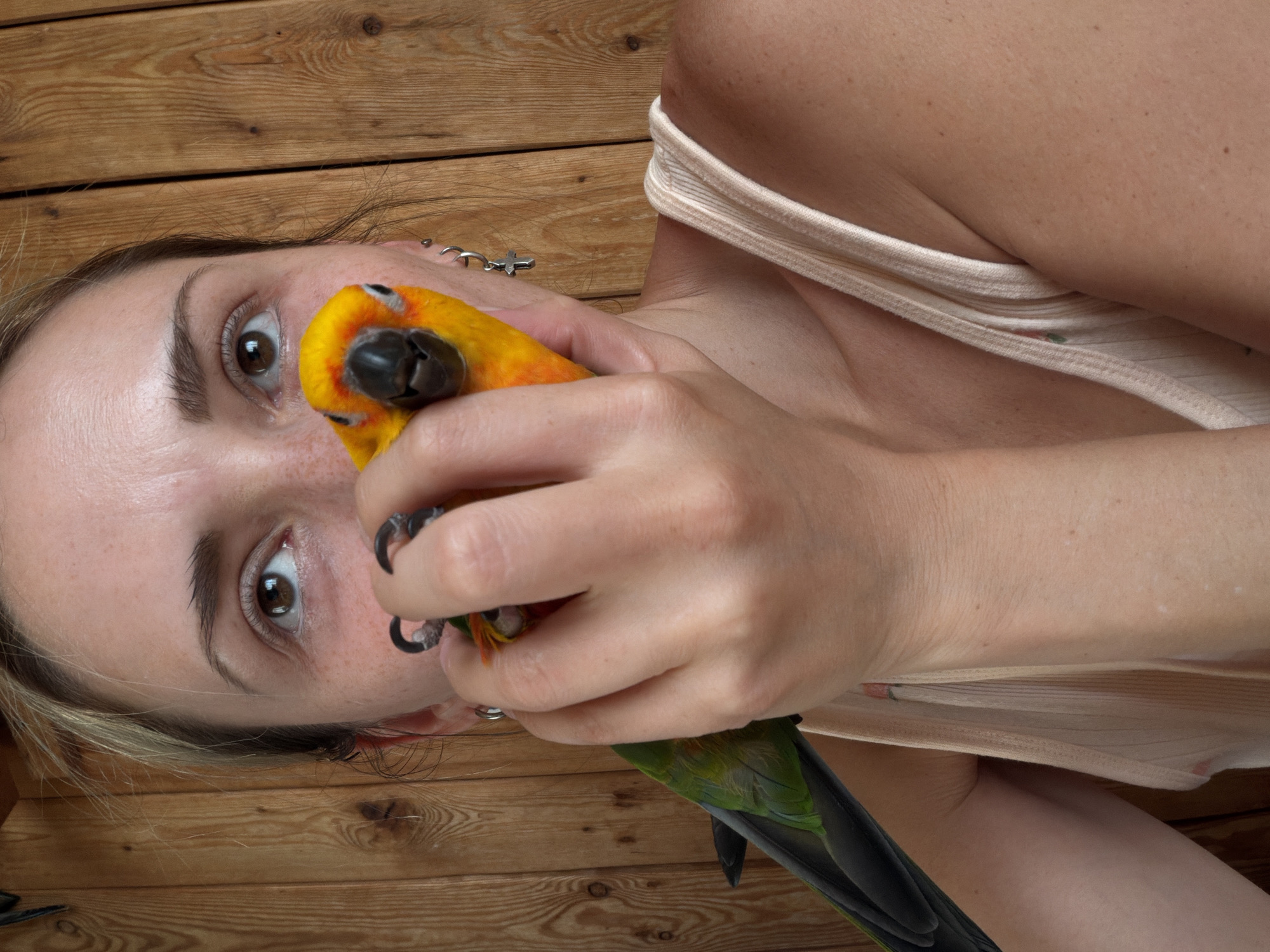 A close-up shot focuses on a woman's face and upper body, with a sun conure parrot perched on her hand, partially obscuring her mouth. The woman has fair skin with freckles and blonde hair pulled back. She is wearing a light-colored tank top. The parrot is bright yellow and orange with green and black accents on its wings and tail. The background consists of wooden paneling. The image does not provide any information about the location in the city of Khimki, Russia.