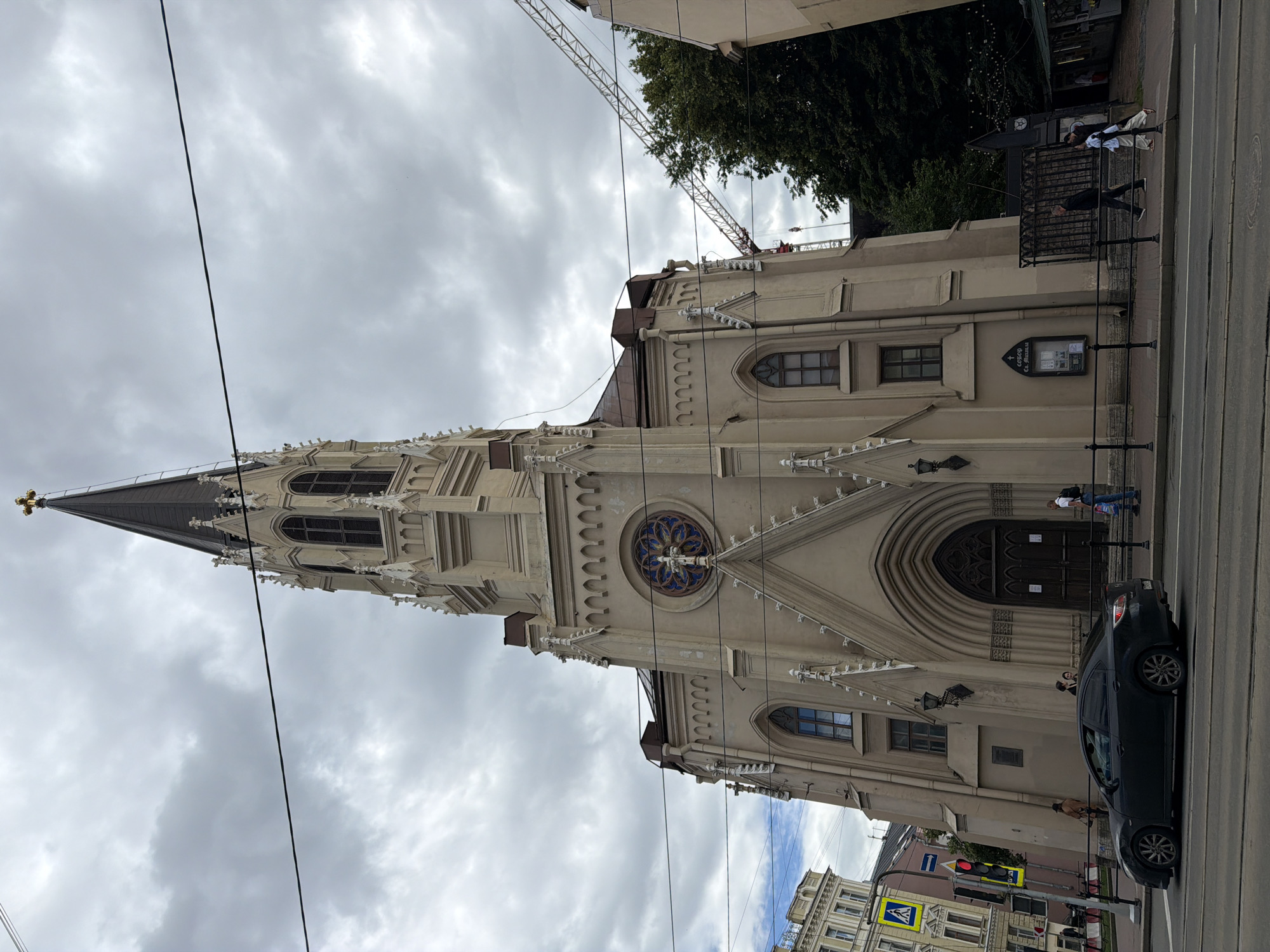 The image shows a light-colored, gothic-style church with a tall spire. The church is located in an urban environment with a street in the foreground and other buildings visible in the background. There are overhead wires crossing the sky. A dark-colored car is parked on the street. Several people are visible walking on the sidewalk in front of the church. The sky is overcast, suggesting cloudy weather. A construction crane is visible in the upper right portion of the image, indicating that some form of construction or renovation may be taking place. The time of day appears to be daytime, given the lighting. The architectural style of the church and the presence of streetlights suggest a European city setting, consistent with the location context of Saint Petersburg, Russia. No text is clearly legible in the image.
