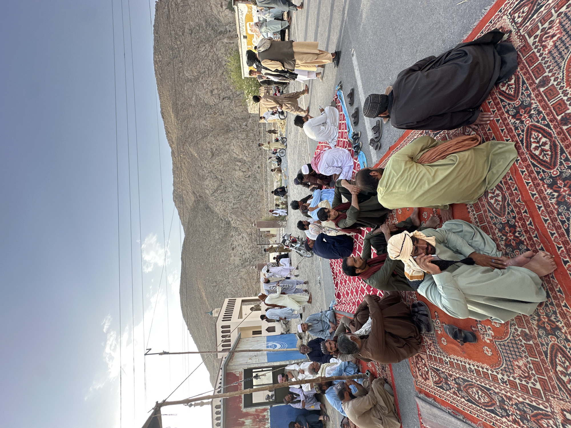 A crowd of men are gathered outdoors, seated on rugs spread on a paved road. In the background, there is a building with an arched entrance and a minaret, possibly a mosque, and a barren, rocky mountain range. Several men are talking amongst themselves, while one man in the foreground is on his phone. There are some motorcycles and a truck visible in the background. The sky is overcast with some clouds. The scene appears to be in Quetta City Tehsil, Pakistan, based on the architectural style and the mountain landscape typical of the region.