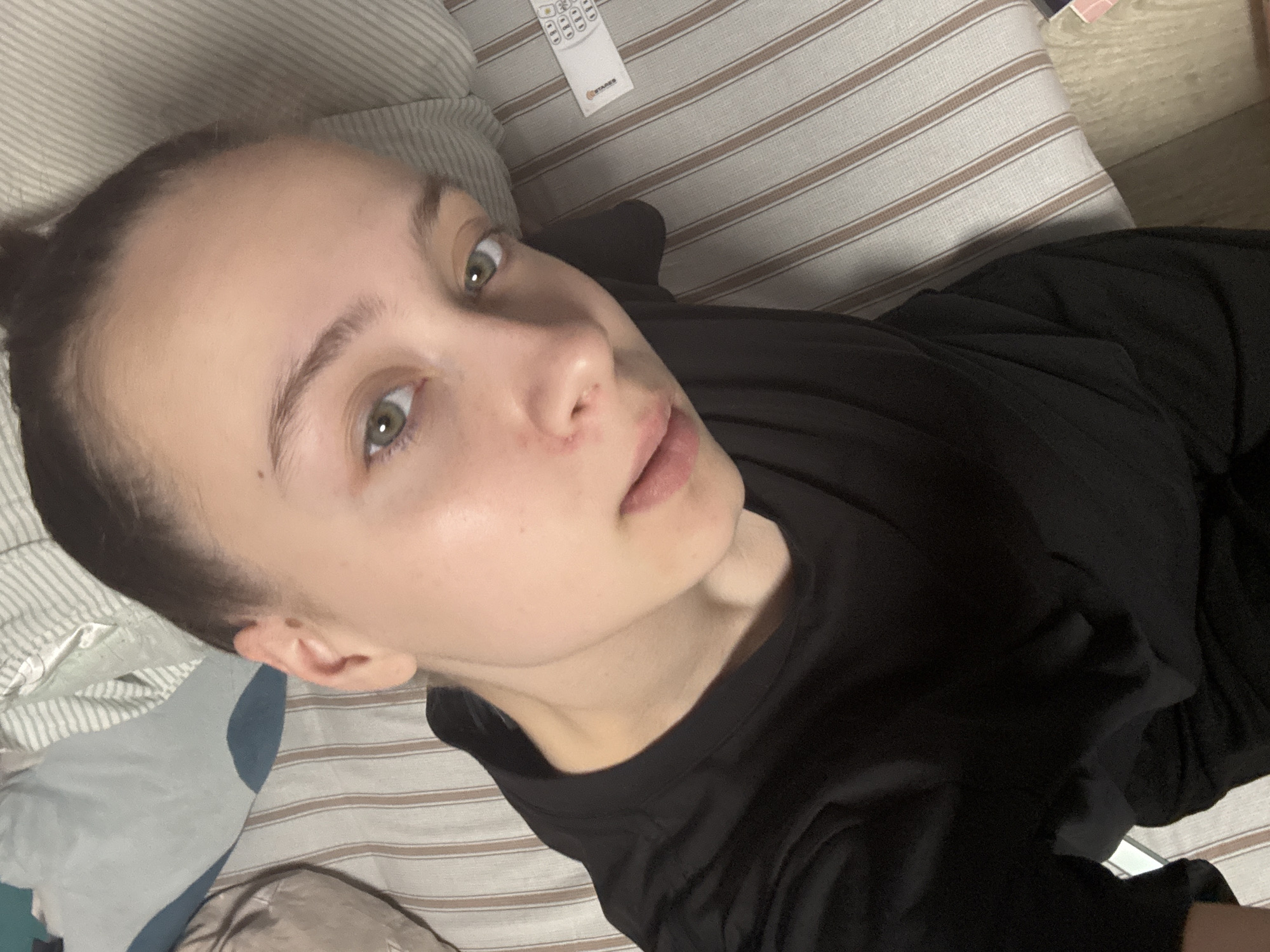 A young woman with fair skin, light green eyes, and dark hair pulled back into a bun, lies on a striped bed or sofa. She wears a black t-shirt and is looking upwards at the camera. A remote control is visible in the upper left corner of the image. The location is presumed to be Прикамский, Russia.