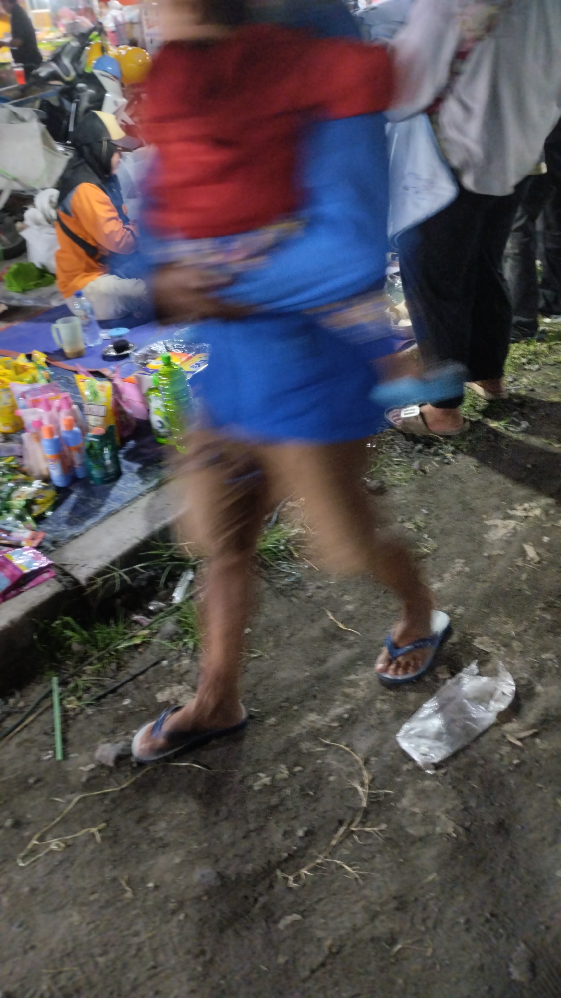 This media file depicts a scene of people and objects at what appears to be an outdoor market or gathering at night in Sambungrejo, Indonesia. The primary focus is on the lower half of a person who is carrying another person, or perhaps a large bag. The person in front is wearing a red top and blue lower garment, while the person being carried is wearing a blue outfit. The legs of the person in front are blurred, indicating movement, and they are wearing flip-flops.
In the background, several other people are visible, some sitting and others standing. One person is wearing an orange jacket and a headscarf. There are also various objects scattered around, including plastic bottles, bags of goods, and what looks like a mug and a plate. The ground appears to be dirt or mud, with some grass and debris.
The overall impression is of a busy, possibly crowded, nighttime event. The lighting is dim, suggesting it is late evening or night. There are no clear indications of the weather, but the ground condition might suggest it has rained or is a generally damp area. The blurriness of the subjects suggests the photo was taken with a fast shutter speed in low light conditions, or there was significant movement during the capture. No visible text is discernible in the image.