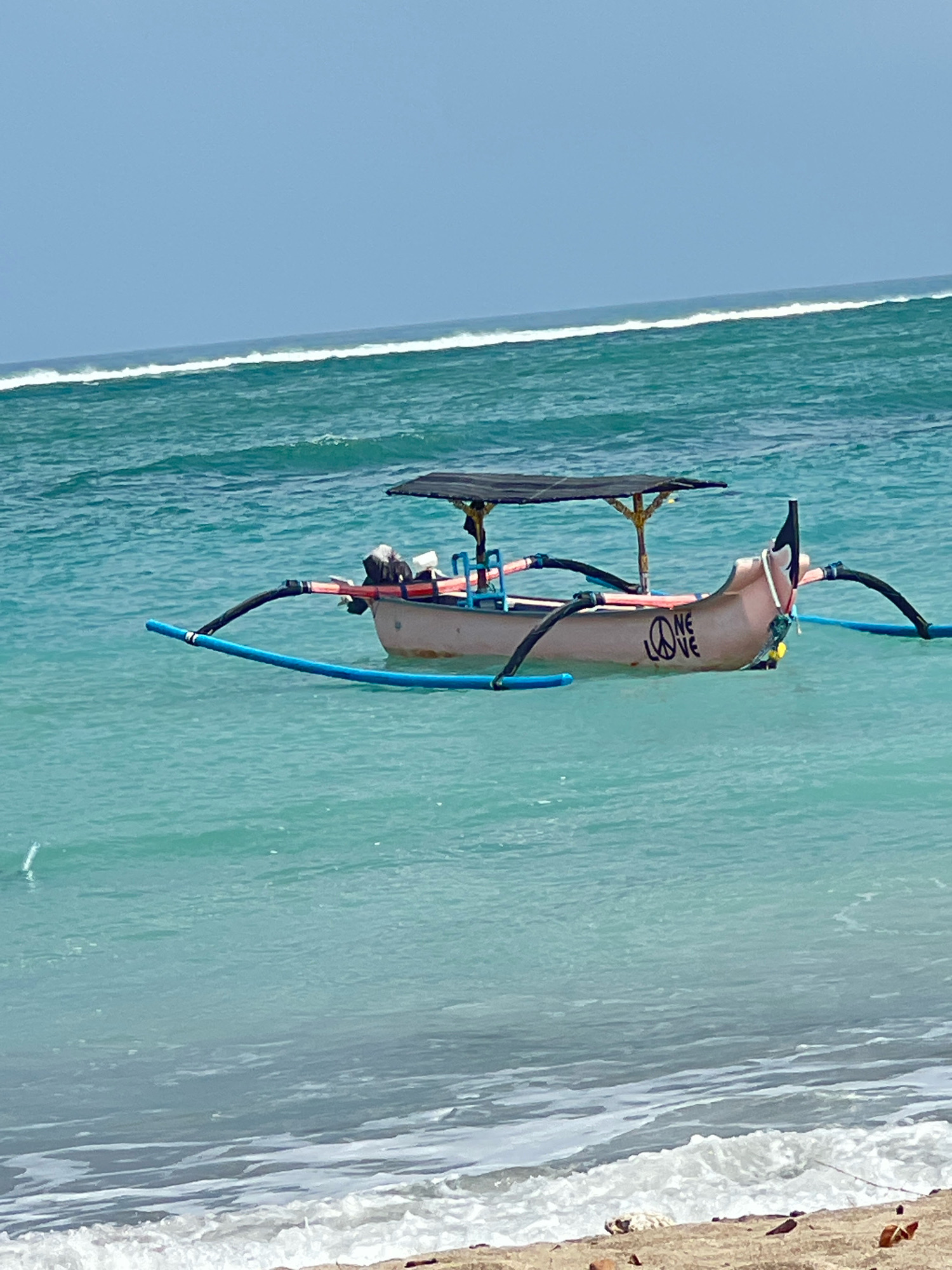 A traditional outrigger fishing boat is anchored in the turquoise waters off the coast of Kuta, Indonesia. The boat, painted a pale pink, has a black canopy supported by bamboo poles, providing shade over the midsection. The words "PEACE LOVE" are stenciled in black on the port side of the hull, with a peace symbol preceding the word "LOVE." Outrigger pontoons, painted bright blue, extend from the sides of the boat to provide stability. The ocean is calm, with gentle waves lapping at the shore in the foreground, where a sandy beach with scattered debris is visible. The sky is a clear, pale blue.