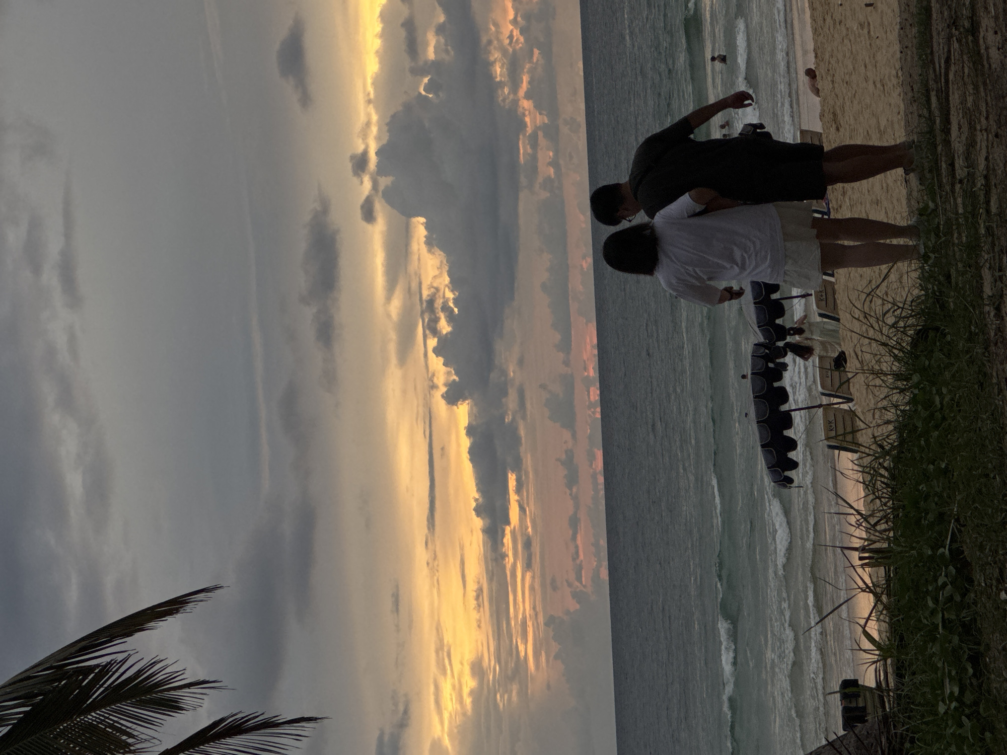 This media file depicts a serene beach scene at sunset in Karon, Thailand. The primary subjects are two individuals, a man and a woman, standing close together on the sandy shore, facing the ocean. The man is wearing a dark t-shirt and shorts, while the woman is in a white top and skirt. They appear to be enjoying the view and each other's company, suggesting a romantic or companionable moment.
The scene is set against a backdrop of a dramatic sky filled with a mix of dark and light clouds, illuminated by the warm, golden hues of the setting sun. The ocean stretches out towards the horizon, with gentle waves lapping at the shore. In the background, other beachgoers can be seen in the distance, and there are lounge chairs and umbrellas set up on the sand, indicating a popular tourist destination. A palm frond is visible in the foreground, adding to the tropical ambiance.
The time of day is clearly sunset, characterized by the soft, diffused light and the rich colors in the sky. The weather appears to be calm, with no strong winds or rain, contributing to the peaceful atmosphere. The presence of lounge chairs and the overall setting strongly suggest a resort or public beach area in a tropical location. The mood conveyed is one of tranquility, beauty, and possibly romance, as the couple takes in the natural spectacle. No visible text is present in the media file.