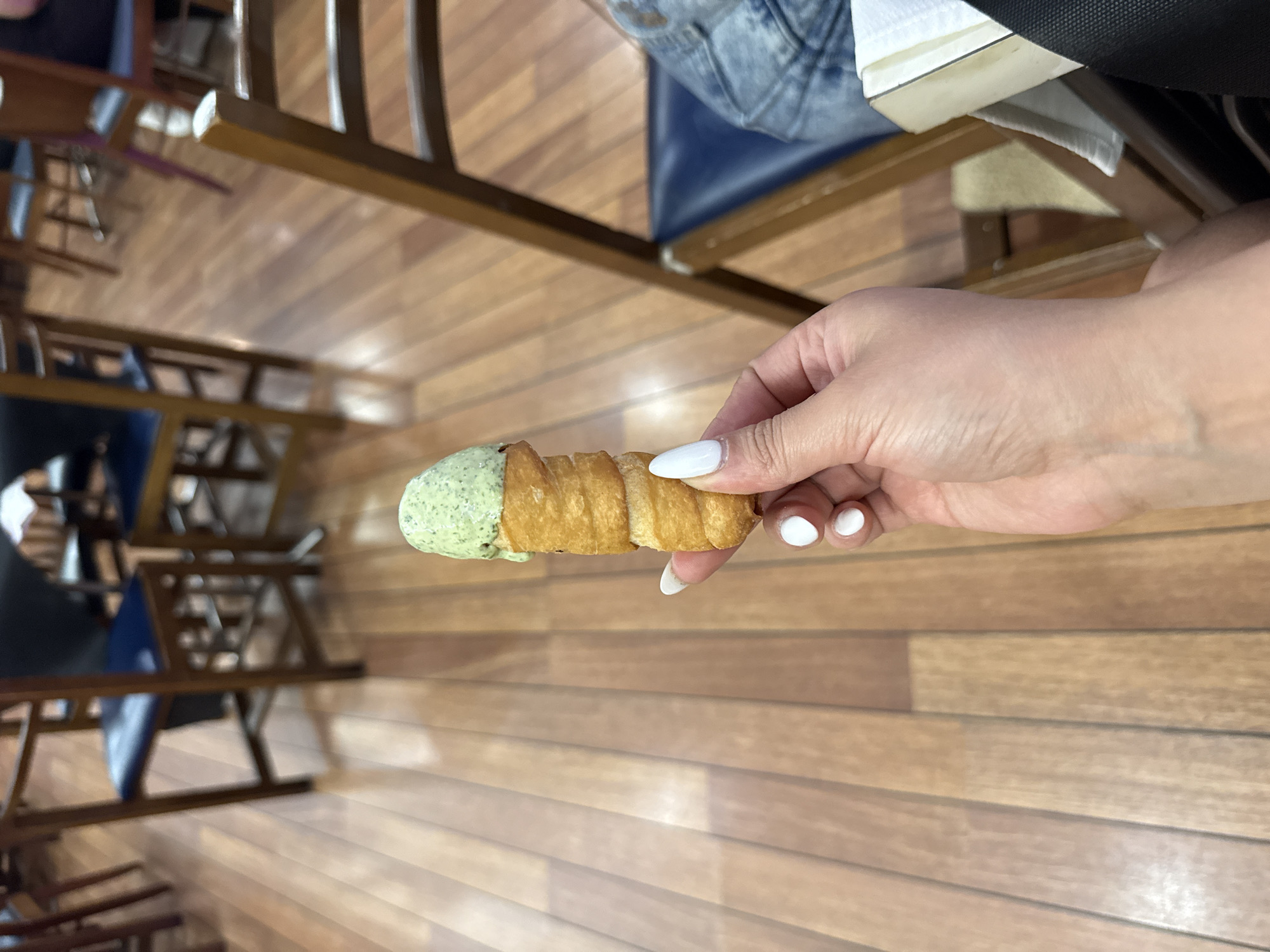 A person's hand with white-painted fingernails holds a cannoli with pale green filling. The cannoli appears to be a fried pastry shell filled with a creamy, textured substance, possibly pistachio ice cream or a similar dessert. The background is a warm-toned wooden floor with a visible grain pattern, suggesting an indoor setting. In the blurred background, there are several wooden chairs with dark blue seats, indicative of a cafe, restaurant, or similar establishment. The lighting suggests it is daytime. There is no visible indication of the location being in Torrejón de Ardoz, Spain.