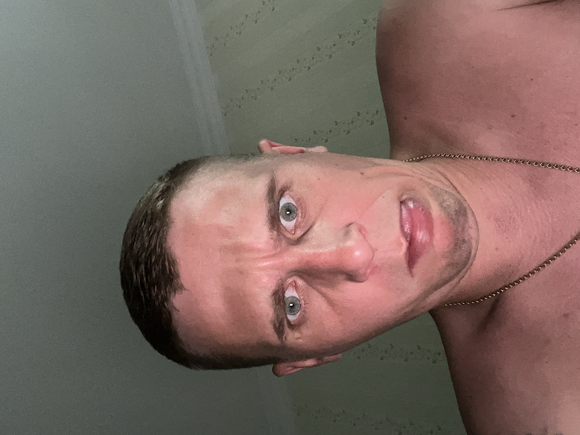 A close-up, low-angle portrait captures an adult male looking directly into the camera. He has short, dark, closely cropped hair and light-colored eyes. His skin appears flushed or reddish, particularly on his face, neck, and shoulders. He displays an exaggerated facial expression with furrowed brows, wide eyes, and his mouth slightly open, revealing the tip of his tongue. He is bare-chested and wears a thin, gold-colored chain necklace. The background indicates an indoor setting with a light green wall featuring a subtle, repeating patterned wallpaper. A white ceiling with a simple molding is visible above. The scene is illuminated by strong overhead artificial lighting, casting a bright, somewhat reddish hue on the subject's skin. While the image itself provides no distinct visual cues to confirm a specific urban location, it is associated with the city of Minsk, Belarus.