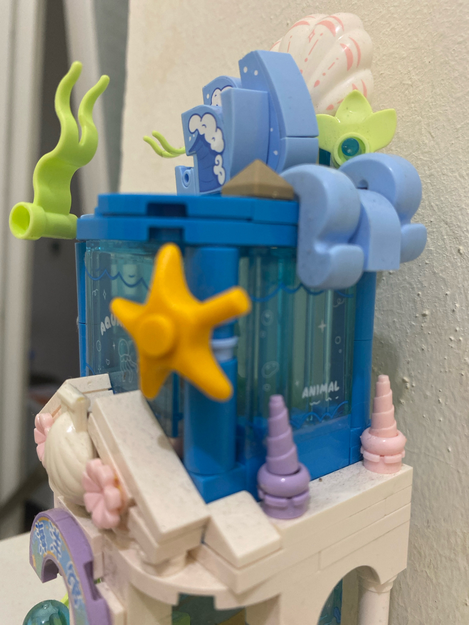 The image is a close-up shot of a colorful, toy building block structure, possibly a LEGO set, with an aquatic or ocean theme. The structure is predominantly blue, light blue, and white, with accents of yellow, green, and purple. Visible elements include a yellow starfish, green wavy seaweed-like pieces, a white seashell, and translucent blue panels with wave and creature graphics. The words "AQUA" and "ANIMAL" are visible on the translucent panels. The structure is built on a white base with arched details and decorative pink flowers. The background is a textured, light-colored wall. There are no people or animals in the image. The location of Kedung Sukodani, Indonesia is not discernible from the image.