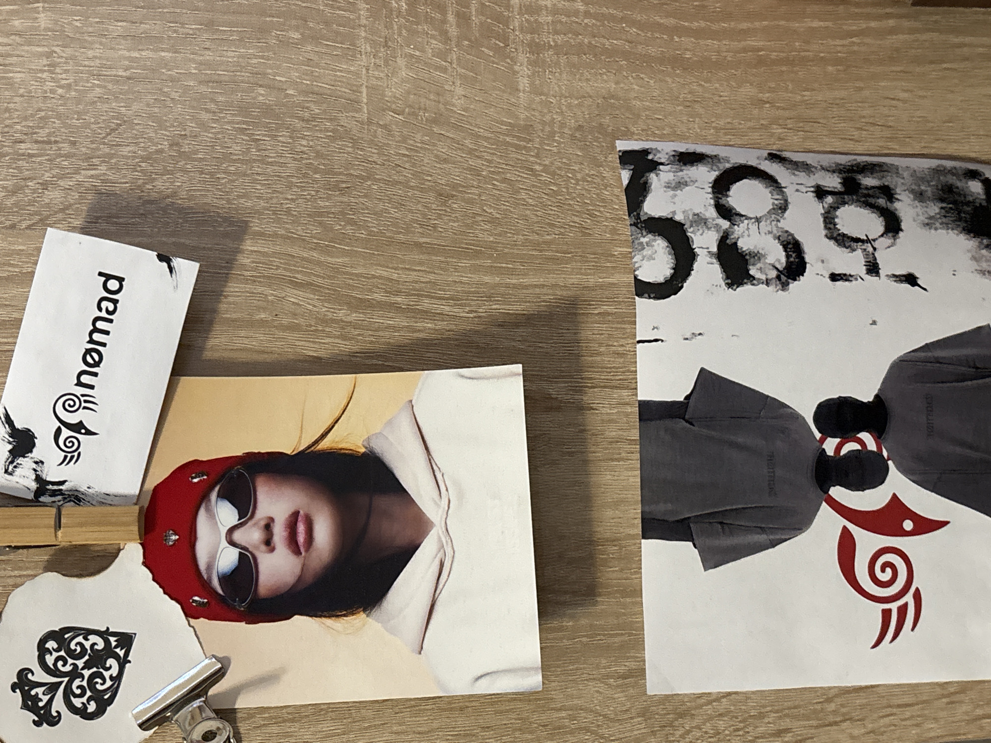 The image shows a collection of items on a wooden surface. A rectangular card with the word "nomad" printed vertically is visible, along with a decorative design. Next to it, there's a photograph of a person wearing sunglasses and a red hat, positioned at an angle. Below these, a paper cutout in the shape of an ace of spades, clipped together with a silver binder clip. To the right, there is another card, featuring the number "588" written in black marker and two human figures wearing grey jackets and red ink design. There is nothing in the image to indicate that this scene is located in the city of Troyan, Bulgaria.