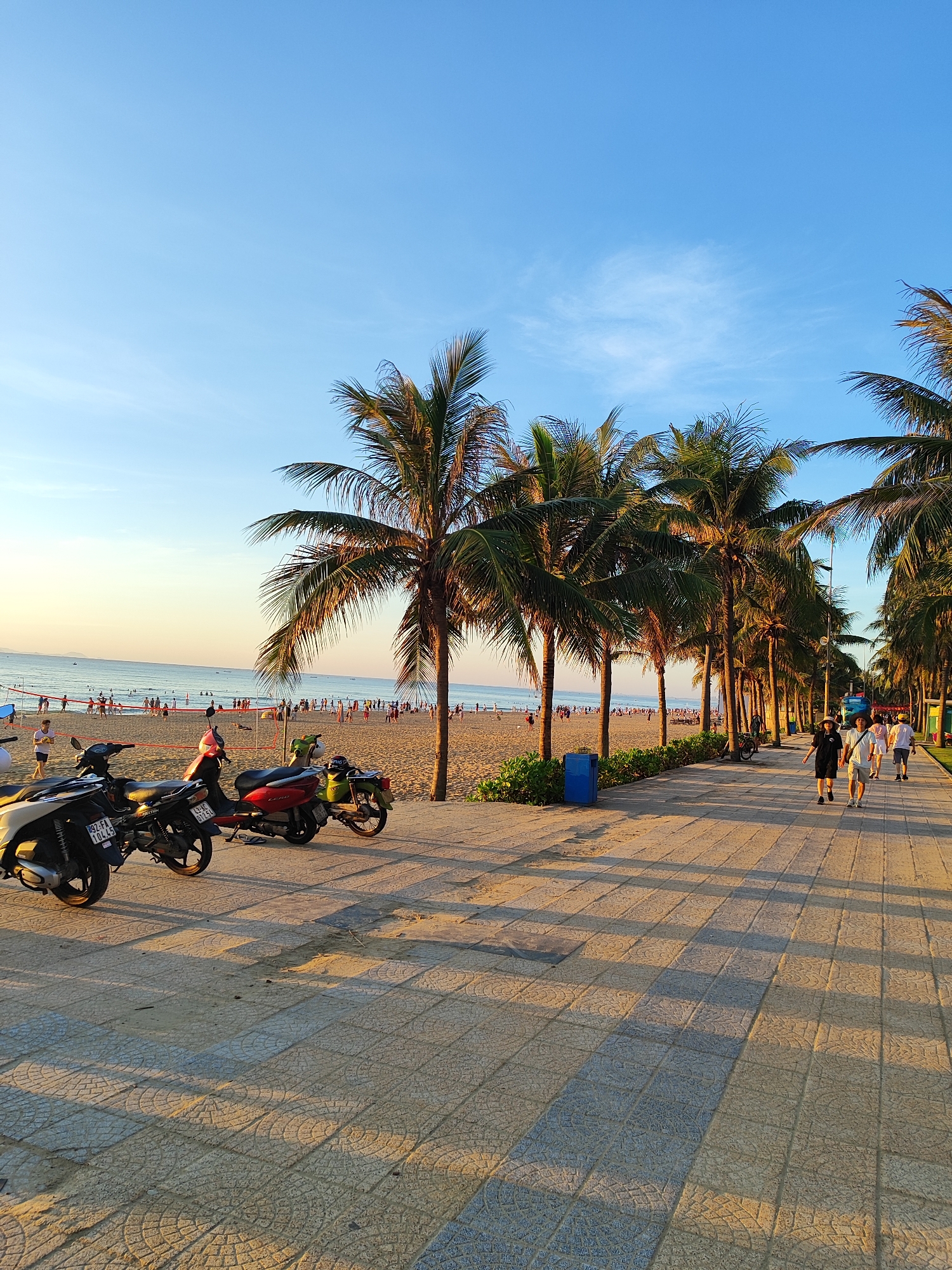 The image shows a beach promenade in Da Nang, Vietnam. In the foreground, several motorbikes are parked on a paved walkway. Beyond the walkway, a sandy beach is populated with many people, and the ocean stretches to the horizon. Palm trees line the promenade, casting shadows on the walkway as the sun begins to set, indicated by the warm golden light. The sky is a clear blue with a few wispy clouds. People are seen walking along the promenade and relaxing on the beach.