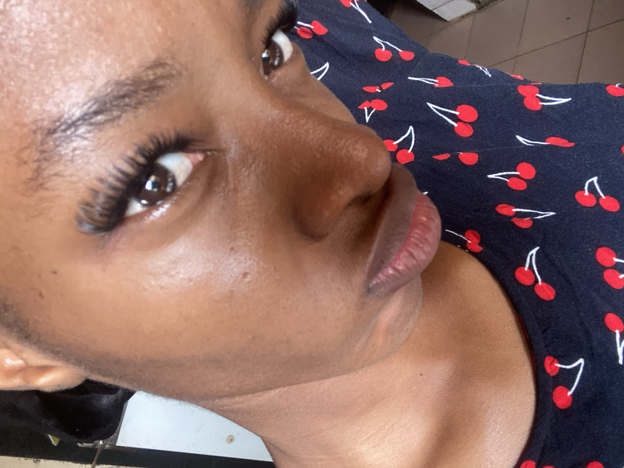 This is a close-up, eye-level portrait of an individual with dark skin, located in the city of Jos, Nigeria. The subject's face dominates the frame, appearing to be tilted slightly downward with direct eye contact made with the viewer. The visible features include prominent dark brown eyes enhanced with false eyelashes on the upper lids, dark eyebrows, and a visible nose bridge and tip. The lower lip is full with a reddish-pink tone. Skin texture is discernible, showing some reflective sheen on the forehead, nose, and cheeks, along with a small dark mark on the upper left cheek/neck area. Dark hair is visible around the left ear and forehead. The individual is wearing a dark-colored garment, likely a top, featuring a repeating pattern of red cherries with white stems, visible around the neckline and extending across the chest and shoulder area. The background elements are minimal: a light-colored vertical surface with a small circular fixture is visible to the left of the subject's neck, and a brown tiled floor is partially visible in the bottom right corner of the frame. A dark, undefined object is also partially visible behind the subject's head on the upper left.