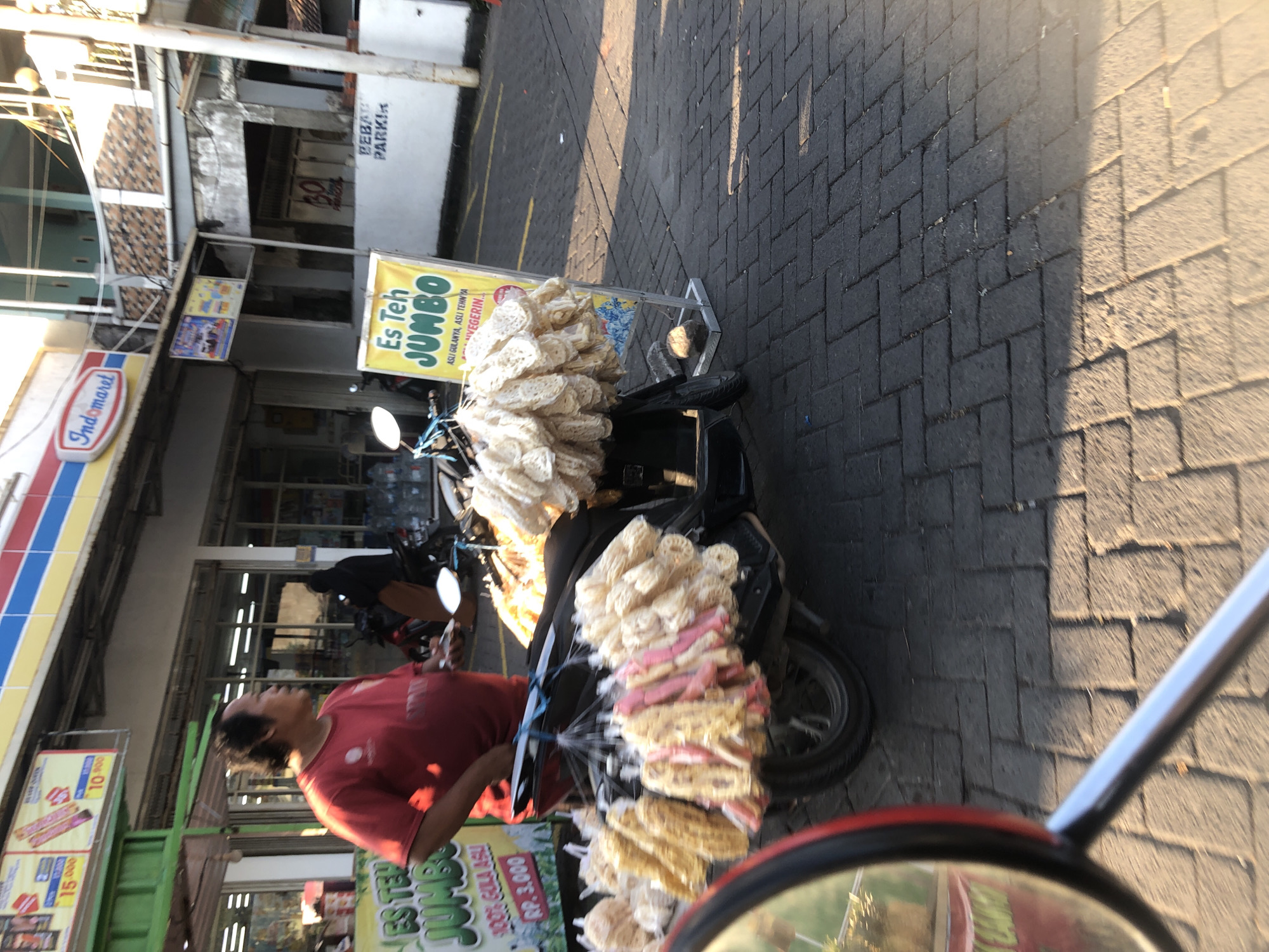 A man wearing a red t-shirt stands next to a motorcycle laden with packaged snacks. The motorcycle is parked on a paved sidewalk in front of a row of storefronts. A sign with "Es Teh Jumbo" is visible, indicating a beverage offering. The scene appears to be in the city of Kalisampurno, Indonesia, as suggested by context. The background shows a building with the "Indomaret" logo, a common convenience store chain in Indonesia. The lighting suggests it is daytime.
