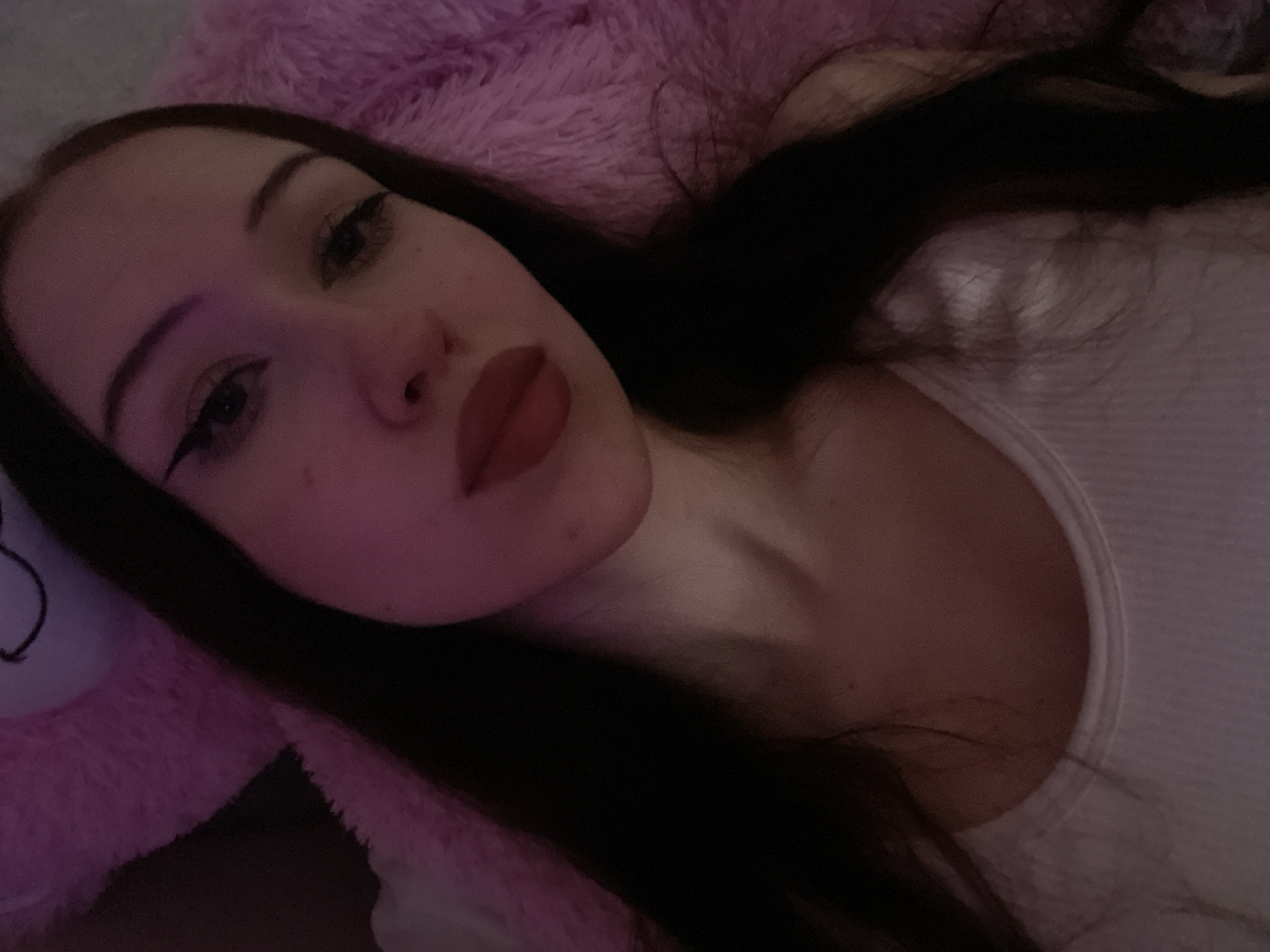 A young woman with long, dark hair is lying down and taking a selfie. She is wearing a white tank top. The background is a pink, fluffy surface, possibly a blanket or stuffed animal, with a hint of purple lighting. The woman has on winged eyeliner and red lipstick. The image is taken from a close-up perspective, focusing on her face and upper chest. There is no discernible location within Votkinsk, Russia.