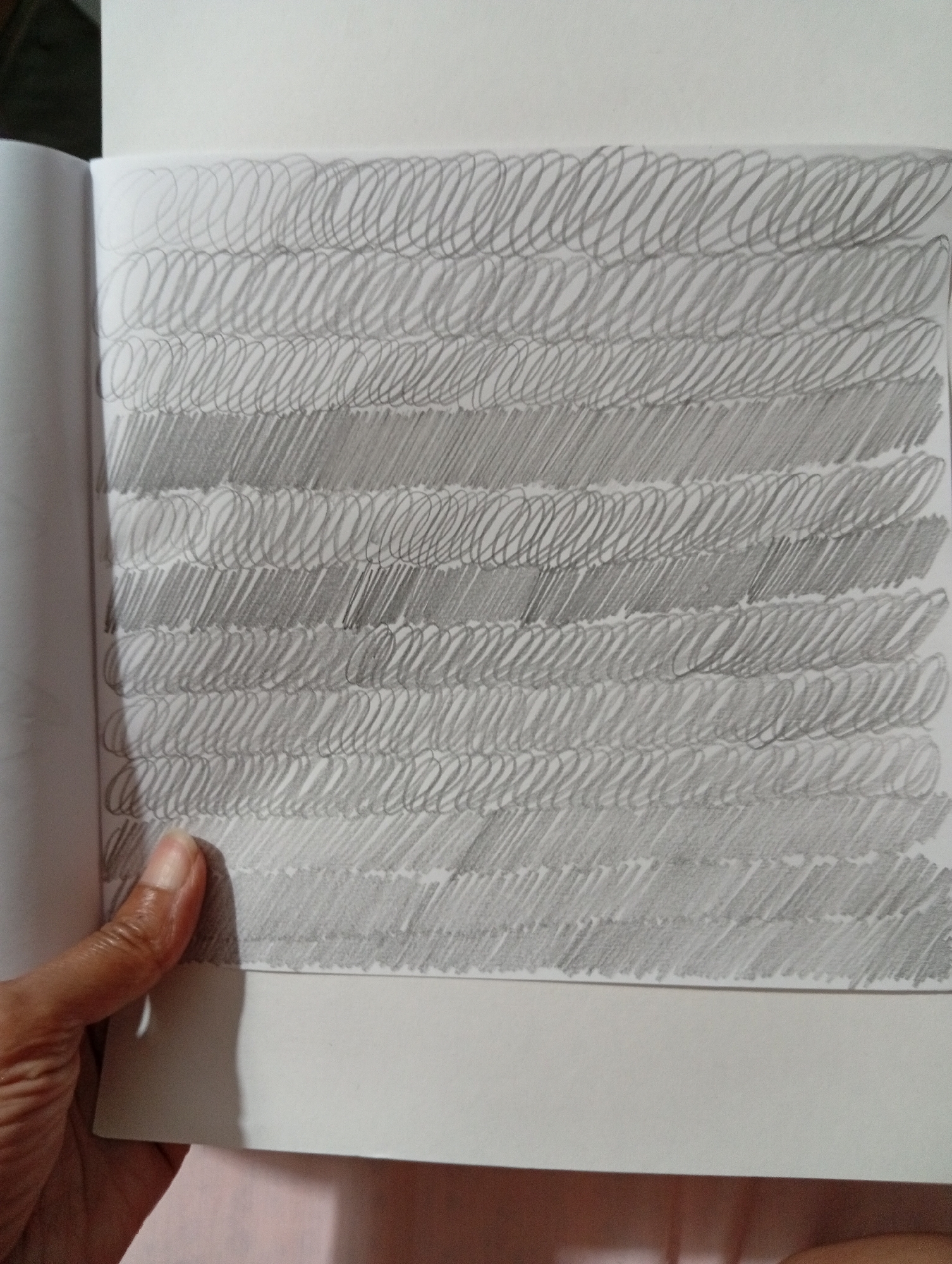 The image displays a close-up view of a sketchpad open to a page filled with horizontal lines of pencil strokes. The strokes vary in density and pattern, creating bands of different shading. Some bands consist of tightly packed diagonal lines, while others feature looser, overlapping circular or oval shapes. A portion of a person's hand and thumb is visible on the left side of the image, holding the sketchpad. The background is a plain, light-colored surface. The scene appears to be indoors, with soft lighting. There is no identifiable information within the image that places it in San Antonio de Los Altos, Venezuela, or any specific city.