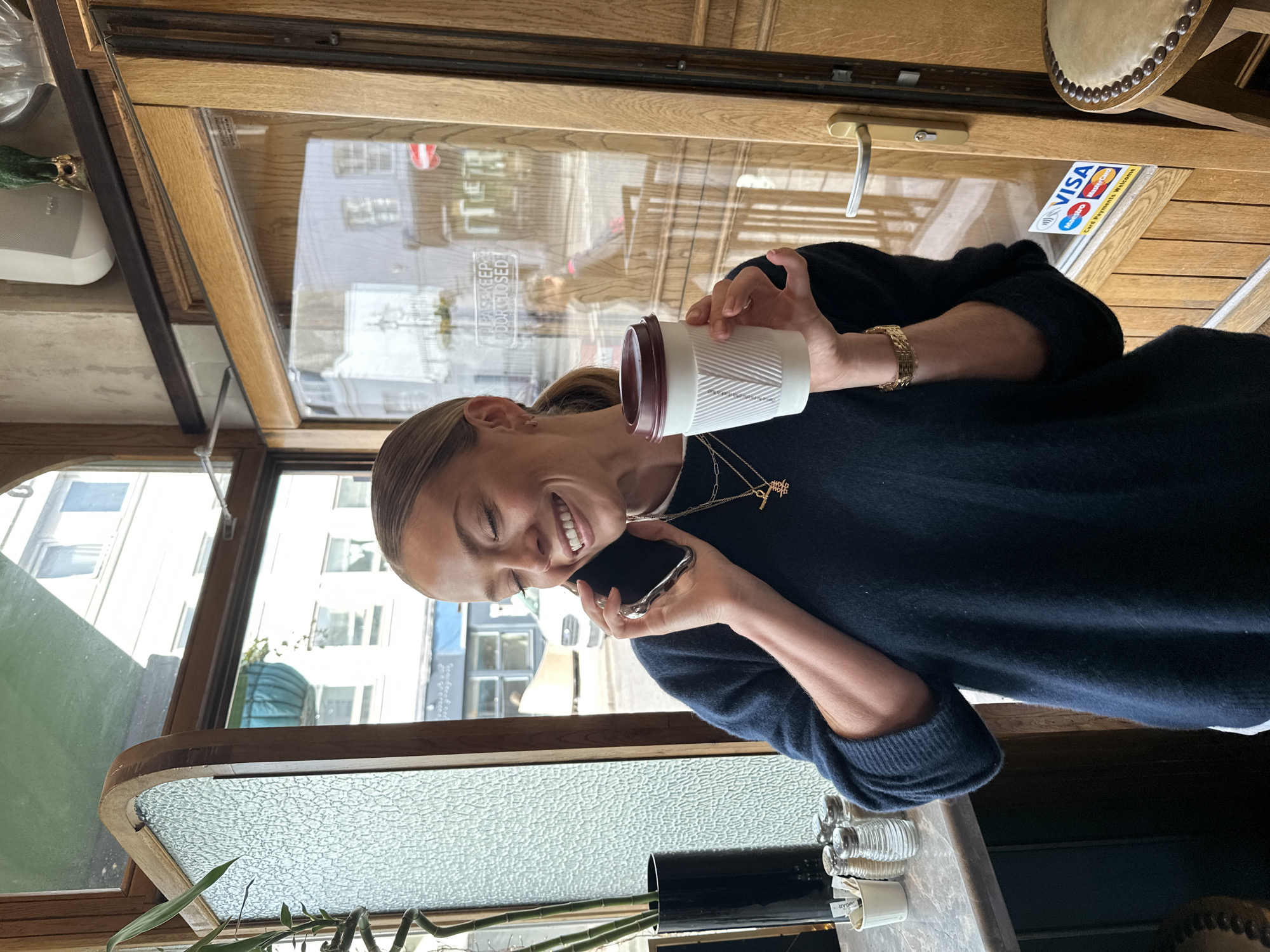 A woman is sitting in what appears to be a cafe or establishment with a tram-like design, possibly in the city of London, United Kingdom. She is wearing a dark blue or black sweater and gold jewelry (necklace and bracelet), holding a white cup with a maroon lid in one hand and a smartphone in the other, pressed to her ear as if on a call. She is smiling and looking towards the camera. Outside the window, buildings can be seen, indicating an urban setting. There is a green plant in a black vase on the table next to the woman. A sticker indicating acceptance of VISA and Mastercard payment is on the window. The establishment has a vintage look with wooden trim and textured glass.