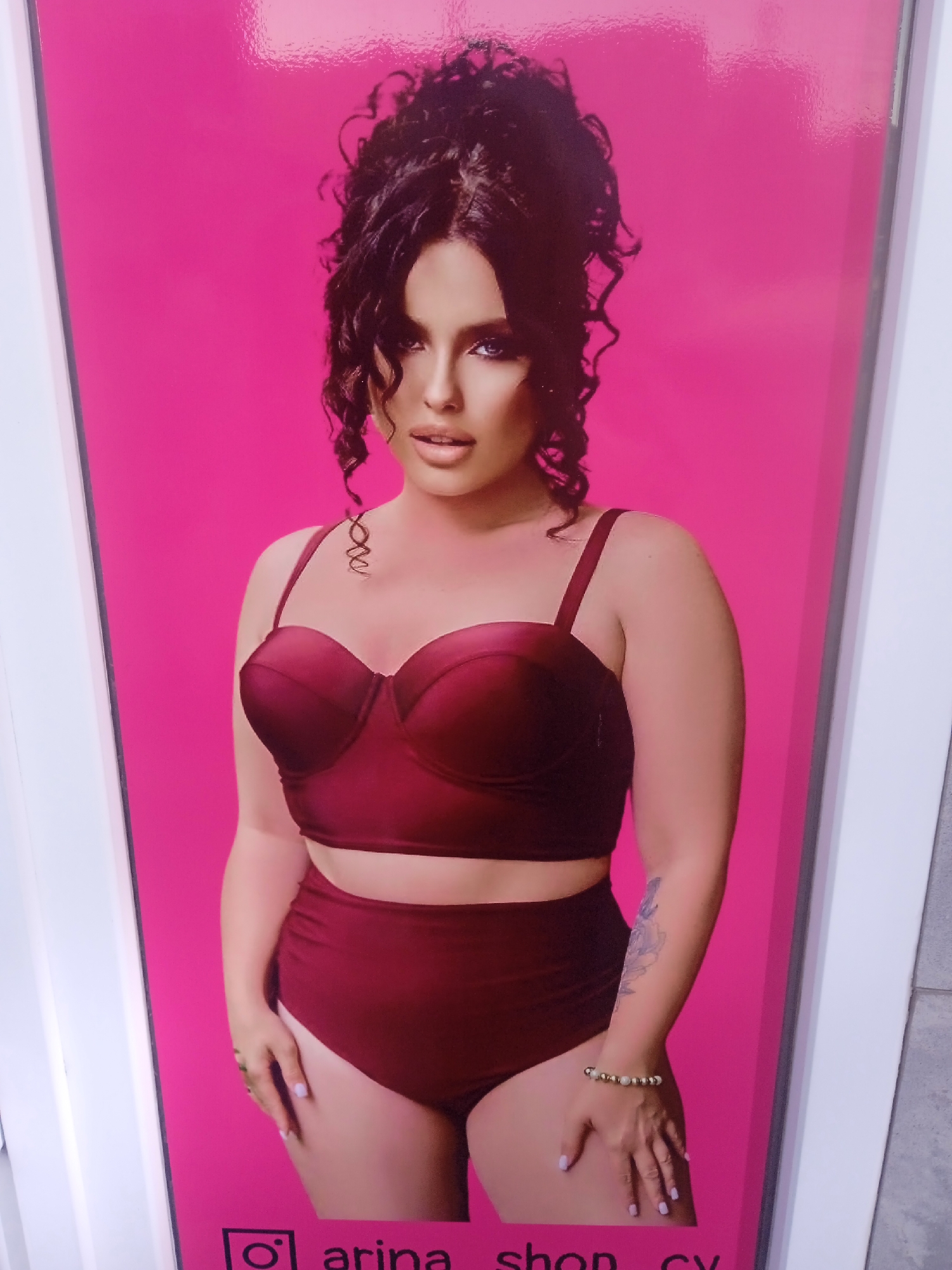 A life-size poster of a woman wearing a burgundy bikini is displayed against a bright pink background. The woman has dark, curly hair styled upwards, and is wearing dark eyeliner and makeup. She is standing and her hands are visible, one resting on her hip. A bracelet is visible on her left wrist, and a tattoo on her left arm. Text with an Instagram logo and the words "arina shop cv" is present at the bottom of the poster. The poster is framed by white borders and is likely displayed in an outdoor or public setting given the presence of a light-colored textured surface visible to the right, possibly a wall or pavement. The location is the city of Chernivtsi, Ukraine.