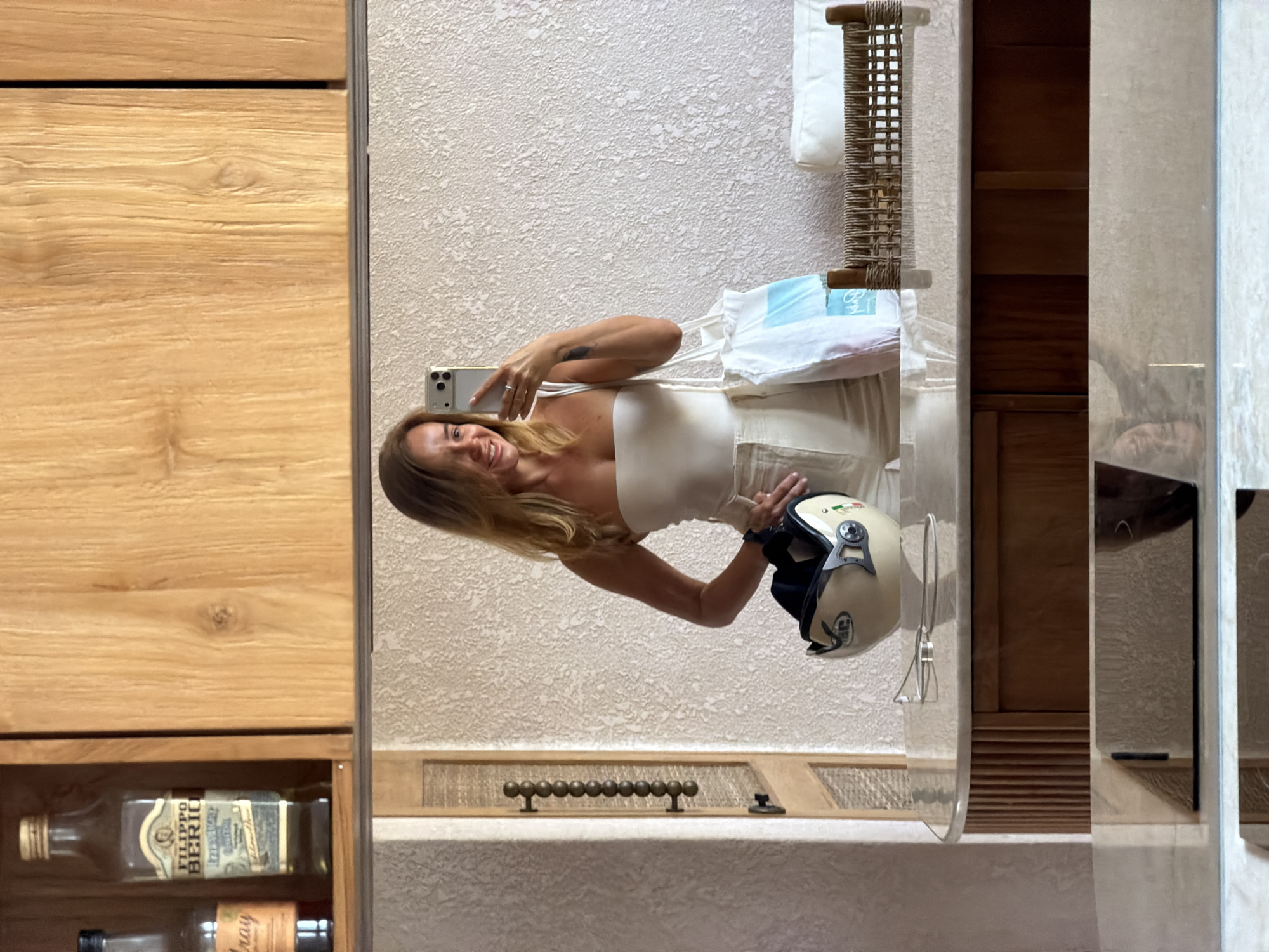 A woman with blonde hair is taking a mirror selfie in a room. She is wearing a white strapless top and light-colored pants. She is holding a smartphone in her right hand and a white tote bag with blue and pink accents is slung over her left shoulder. A beige helmet is resting on a marble countertop in front of her. In the background, there are wooden cabinets and textured walls. The scene appears to be indoors, possibly in a hotel room or a well-decorated home.