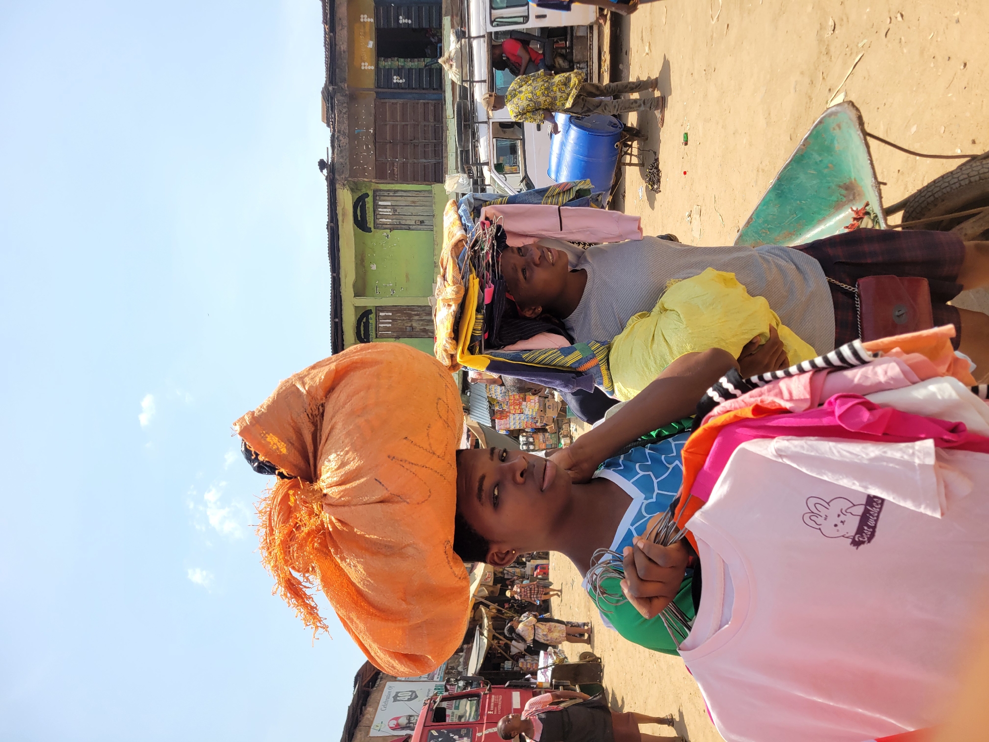 An outdoor, eye-level shot captures a vibrant market scene in Onitsha, Nigeria, under a bright, clear blue sky. The image focuses on two young women, likely street vendors, expertly carrying merchandise on their heads.
In the foreground on the left, a young woman faces forward, carrying a large, orange, burlap-like sack on her head, its fringed edges visible. In her arms, she holds a bundle of metal clothes hangers along with several brightly colored t-shirts in shades of pink, green, orange, and white. One pink t-shirt clearly displays a graphic of a bunny and the text "Best wishes." She wears a blue and green patterned top.
To her right, slightly behind, another young woman is visible, also carrying a substantial stack of folded clothes on her head. She wears a grey t-shirt, partly covered by a yellow garment. Both women maintain serious or neutral expressions as they navigate the market.
The background reveals a bustling environment with old, somewhat dilapidated two-story buildings featuring faded green and yellow paint, corrugated iron roofs, and metal security doors or boarded windows. Several vehicles, including white vans and a red truck, are parked or moving on the dusty, unpaved ground. Other people, some appearing to be vendors or shoppers, are scattered throughout the scene. A blue barrel and a green cart are also visible in the mid-ground. The bright sunlight suggests it is midday or early afternoon.