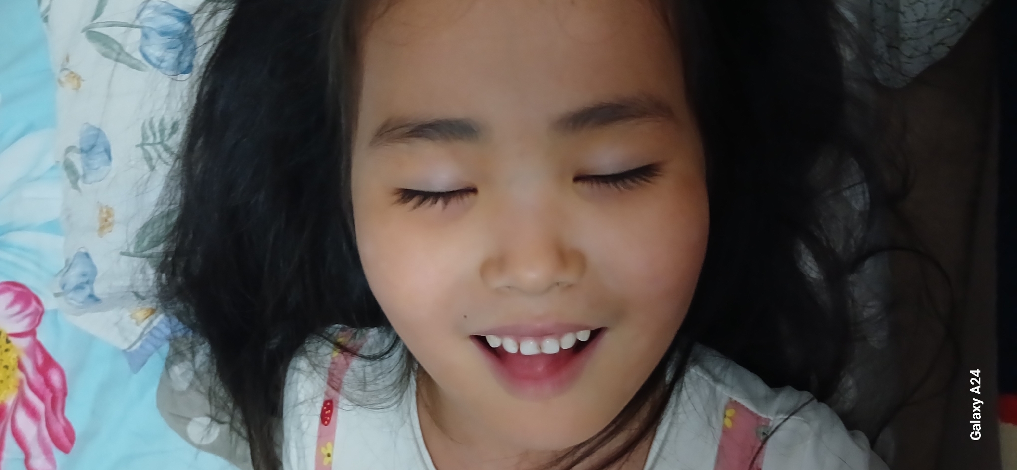 A close-up, rotated view shows a young child with dark hair lying down. The child's eyes are closed, and their mouth is slightly open, revealing small white teeth. The child is wearing a white shirt with a pink and yellow floral pattern. The background appears to be a bed with a light blue pillow that has a floral pattern of blue flowers and leaves. The bottom left corner of the image shows text that reads "Galaxy A24". The image is taken indoors and the lighting suggests it may be daytime. There is no identifiable information about the specific location within Bishkek City, Kyrgyzstan.