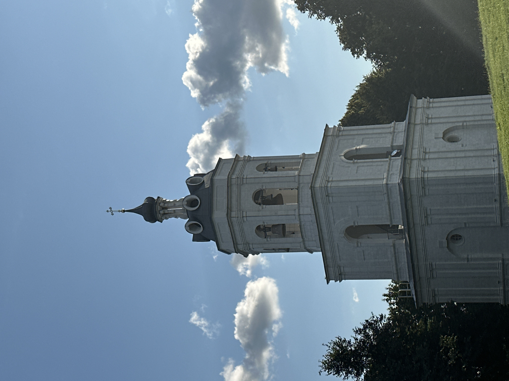 The image shows the bell tower of the Church of the Nativity of the Blessed Virgin Mary in Dubrovitsy, Russia. The bell tower is white and has multiple tiers, with bells visible in the upper section. It is topped with a dark spire and a cross. The sky above is blue with scattered white and grey clouds. Lush green trees frame the sides of the bell tower, and a grassy slope is visible in the foreground. The lighting suggests it is daytime, possibly late afternoon, with some sunlight casting rays across the scene. There are no people or animals present in the image.