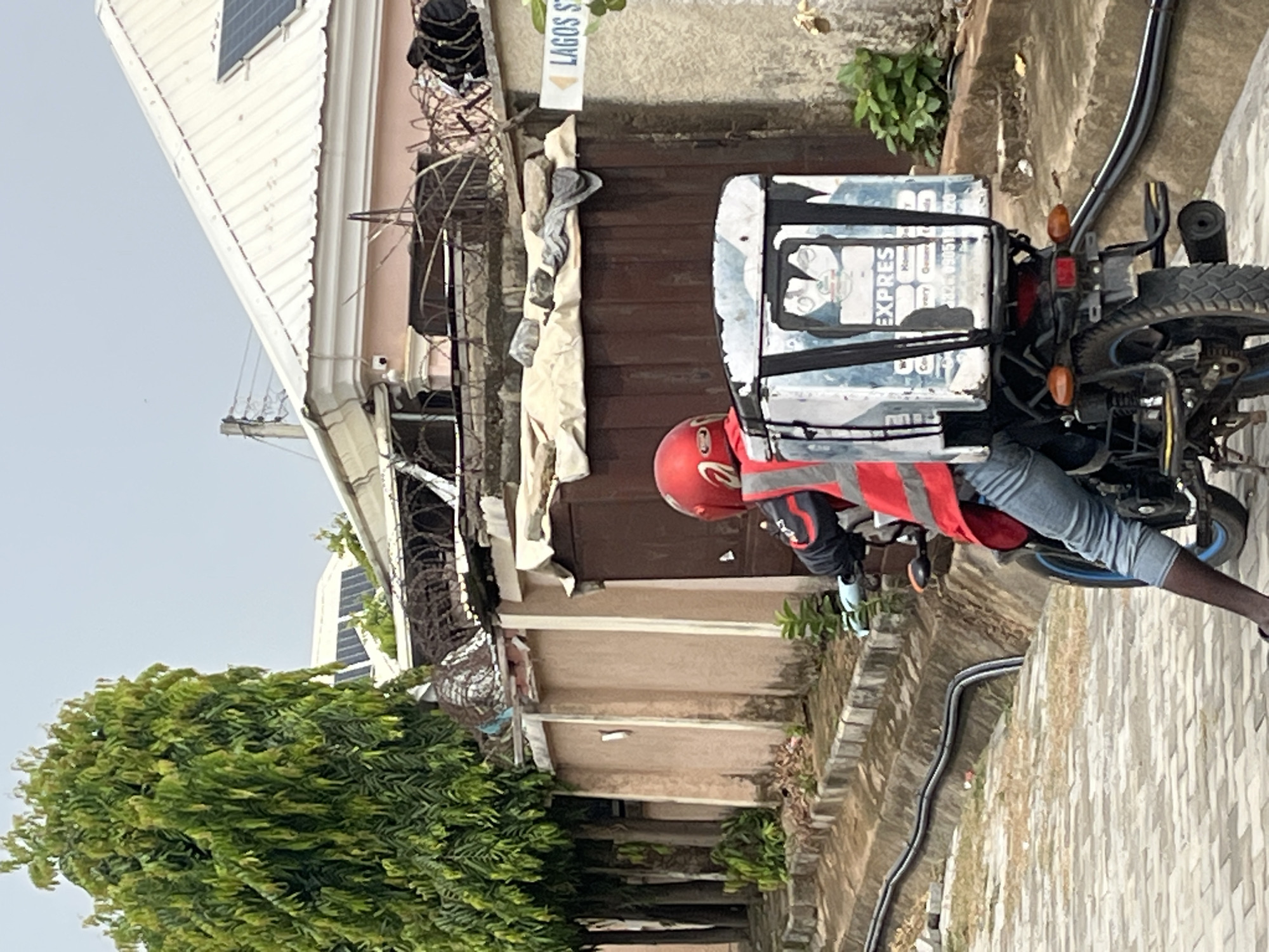 A motorcyclist wearing a red helmet and reflective vest is parked on a street. The motorcycle has a delivery box attached to the back. The Lagos State sign is visible on a building in the background. The environment suggests an urban setting. The location of the photograph is in Abuja, Nigeria.