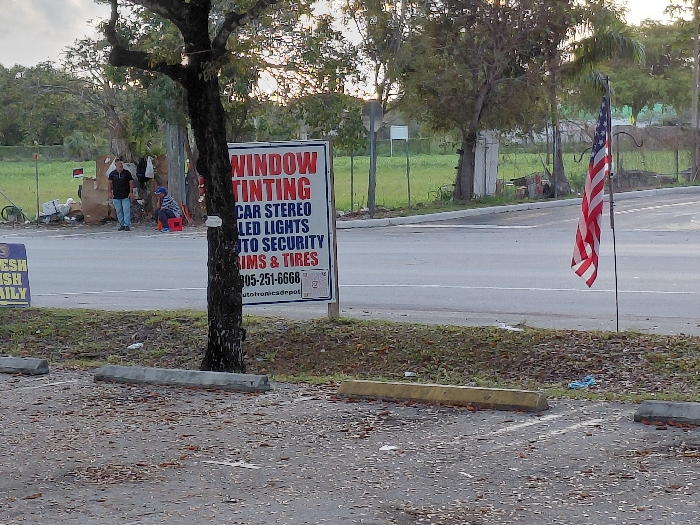 The image depicts an outdoor scene in Cutler Bay, United States of America. In the foreground, a dark, textured ground, likely asphalt or gravel, is covered with scattered brown leaves. Two concrete parking barriers are visible; one gray, and one yellow. A partial blue and yellow sign, reading "RESH" and "SH DAILY," is in the lower left corner.
A grassy median runs horizontally across the midground, separating the foreground from a two-lane asphalt road. A tree with dark, rough bark stands within this median, slightly left of center. To the right of the tree, a prominent white rectangular sign displays services in red and blue text: "WINDOW TINTING," "CAR STEREO," "LED LIGHTS," "AUTO SECURITY," and "RIMS & TIRES." A phone number "305-251-6668" and a website "autotronicsdrpot" are also visible, along with two QR codes. Further right in the median, an American flag is displayed vertically, hanging from a dark metal shepherd's hook.
Across the asphalt road, in the background, two individuals are visible near a cluttered area consisting of cardboard boxes, white plastic bags, and other debris. One person stands, wearing a dark shirt and blue jeans, facing right. Another person is seated on a red stool or bucket, wearing a blue cap and a striped shirt, facing left. A dark bicycle leans against the accumulated items. A wire fence runs behind this area, separating it from a greener, undeveloped lot. Various trees and shrubs populate the background, beneath an overcast or twilight sky.