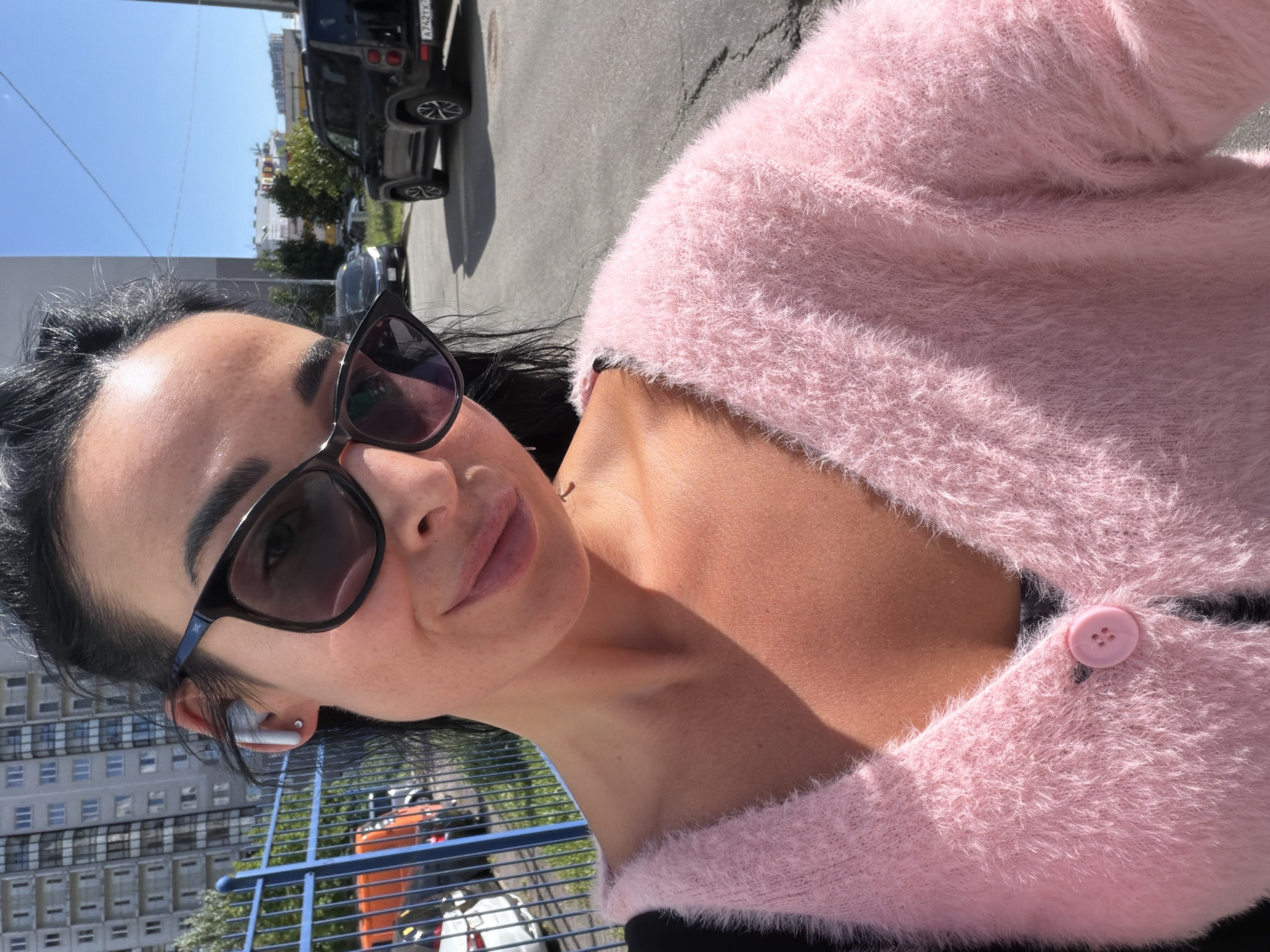 A woman with dark hair and sunglasses is taking a selfie outdoors. She is wearing a fuzzy pink sweater with a single pink button. In the background, there is a tall residential building, a blue fence, trees, and several cars parked on an asphalt road. One car's license plate is visible and appears to be from Russia. The sky is clear and blue.