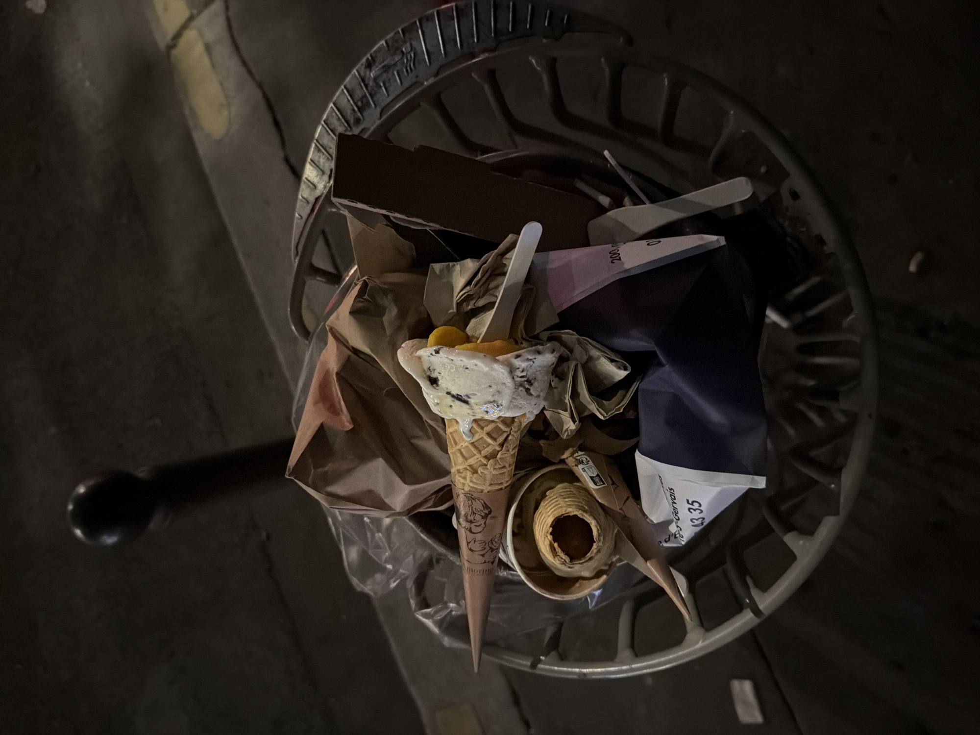 An overhead, close-up shot shows an overflowing, light-gray, metal trash can positioned on a city street in Paris, France. The can is filled with discarded items including an ice cream cone, paper bags, a white plastic spoon, cardboard, and a receipt showing "Saint-Gervais" and "43 35". The ice cream cone, topped with white and dark ice cream and yellow toppings, is prominently placed. Part of a black pole is visible to the left of the trash can. The ground around the trash can is dark and appears to be asphalt. The lighting suggests it is nighttime.