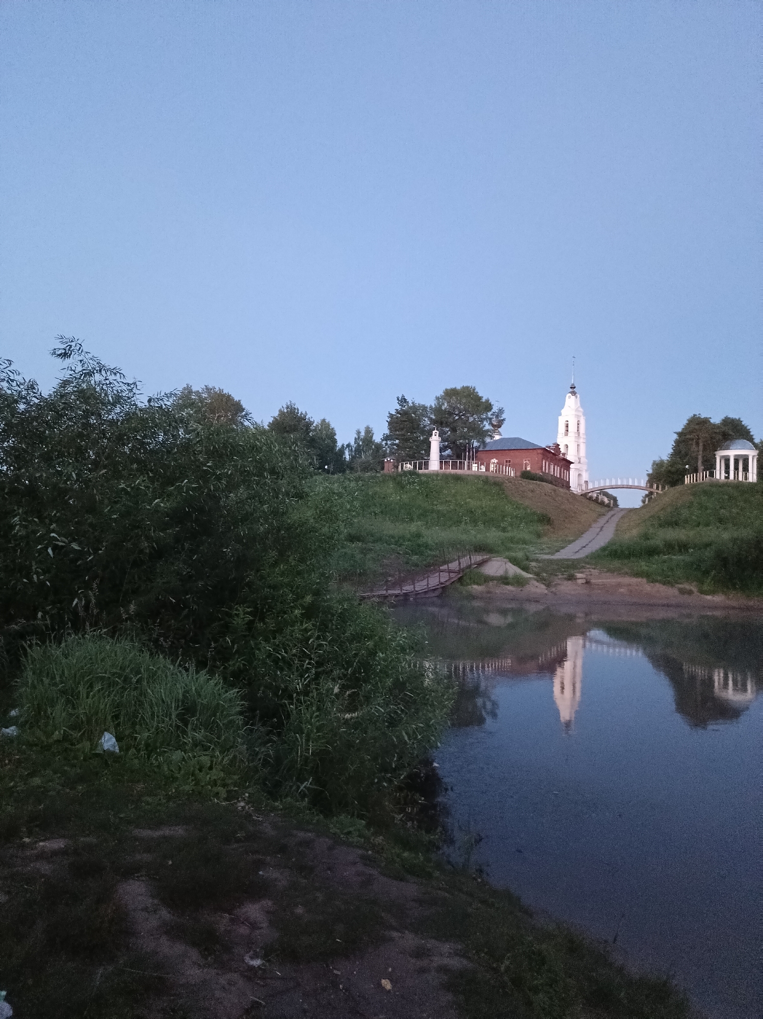 The image shows a serene landscape scene near the city of Buy, Russia, likely during twilight. The foreground features a body of water with a smooth, dark surface reflecting the sky and structures on the opposite bank. To the left, lush green grass and dense bushes dominate the edge of the water, with a white plastic bag visible amongst the foliage.
Across the water, a gently sloping hill is covered in vibrant green grass. Atop the hill stands a red brick building with a white steeple, characteristic of Orthodox churches. Adjacent to the church, a white gazebo with a domed roof is also visible. A narrow, arched pedestrian bridge spans between the two structures on the hill. A path leads down from the bridge towards the water, and a rustic wooden bridge extends from the grassy bank towards the water. The sky above is a clear, pale blue, transitioning into a darker hue towards the horizon. The overall atmosphere is tranquil and picturesque, highlighting the architectural beauty of the church and its natural surroundings.