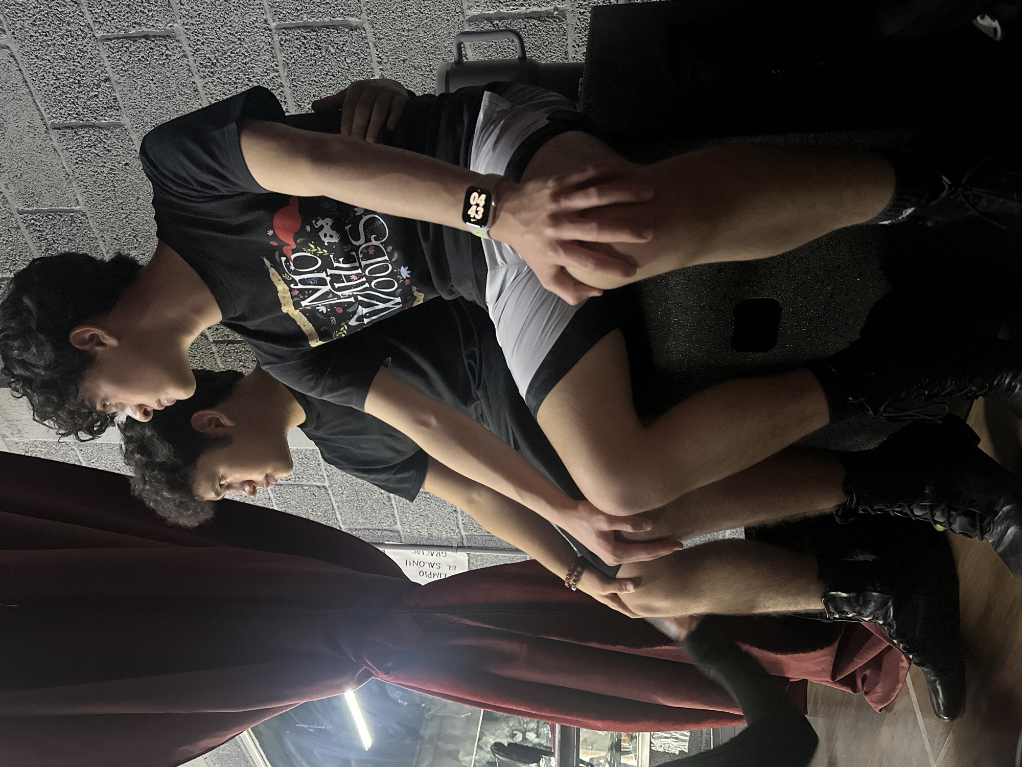 Two young males are seated on a black speaker. The male on the left has dark curly hair and is wearing a black t-shirt and black boots. The male on the right has dark curly hair and is wearing a black t-shirt with a graphic, grey athletic shorts, and black socks with white accents. He is also wearing a smartwatch. The background consists of a textured grey brick wall and a dark red curtain. A sign with Spanish text is partially visible on the wall behind the curtain. The lighting suggests an indoor environment. The location is San Bernardino Tlaxcalancingo, Mexico.