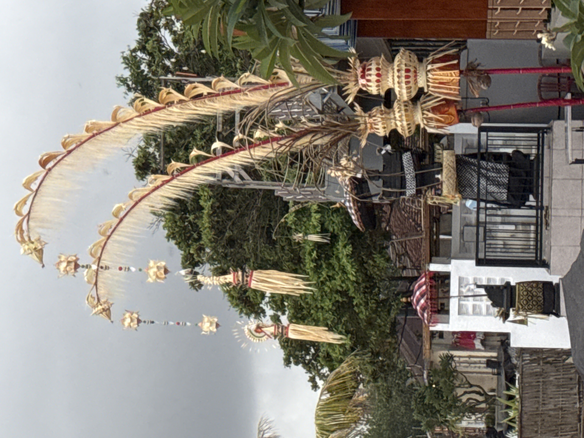 The image depicts an outdoor scene in Kuta, Indonesia, featuring traditional Balinese decorations. In the foreground, two tall, ornate structures made of woven material, likely palm leaves, are prominently displayed. These structures are topped with layered, basket-like elements in shades of gold and red. Hanging from decorative arches made of dried leaves and red sticks are several smaller, intricately crafted ornaments, some resembling flowers or stylized figures, strung together with colorful beads. These arches are tall and curve upwards, framing the overcast sky.
Behind the decorative structures, various buildings and natural elements are visible. A white building with a red and white striped umbrella on top is situated to the left. A dark, ornate statue or offering stand is placed in front of this building. Further in the background, lush green trees with dense foliage are present. Some of the buildings have tiled roofs, typical of the region. A black metal fence runs across the mid-ground, partially obscuring the view of the buildings behind it. Wooden fencing is visible on the far left. The overall atmosphere is one of a festive or ceremonial occasion, with the decorations suggesting a cultural event or religious observance in Kuta. No people are visible in the frame. The lighting suggests it is daytime, but the sky is cloudy.