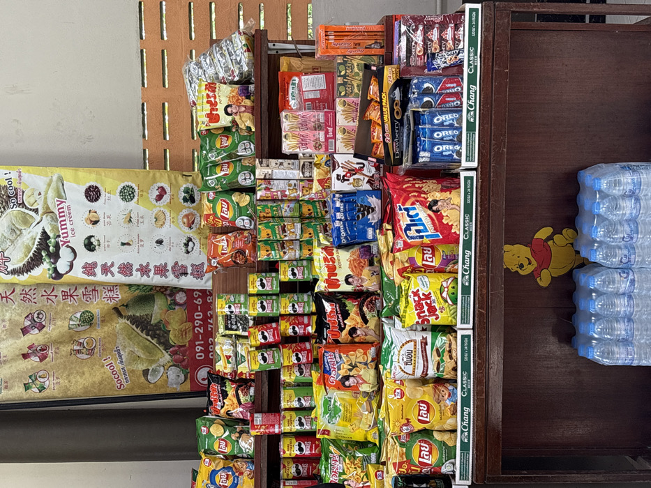 This image captures a multi-tiered retail snack and beverage display unit located in a facility in Takua Thung, Thailand. The primary subject is a dark brown wooden display stand laden with various packaged food items.
Above the display, two large vertical banners are affixed to a wall. The left banner features illustrations of tropical fruits like durian, mangosteen, and coconut, promoting "Socool!" products, with Thai and Chinese text and a phone number. The right banner, light yellow, advertises "Yummy ice cream" and "天然水果雪糕" (Natural Fruit Ice Cream), also displaying various fruit illustrations. The wall behind the banners is painted light grey on the left and transitions to a light orange, horizontally patterned brick wall on the right.
The wooden display stand presents merchandise across multiple shelves. The upper shelves are heavily stocked with numerous bags of potato chips, predominantly Lay's brand in various flavors, some featuring a Thai celebrity. Rows of Pringles potato crisp cans in diverse colors and flavors are also prominently displayed. Other snack items include bags of "Snack Jack," "Oreo" cookies, "Glico Pocky" stick biscuits, and "Dewberry" branded items, arranged in a dense retail configuration. Some snack bags are positioned within white plastic trays bearing the "Chang CLASSIC beer" logo and text indicating "120 ml / 24 cans."
At the base of the wooden display stand, several multi-packs of bottled drinking water are stacked. A yellow, flat cutout of Winnie the Pooh is affixed to the front, lower section of the wooden stand, positioned to the left of the water bottles. No individuals are visible in the scene. The overall lighting suggests an indoor or sheltered outdoor environment.