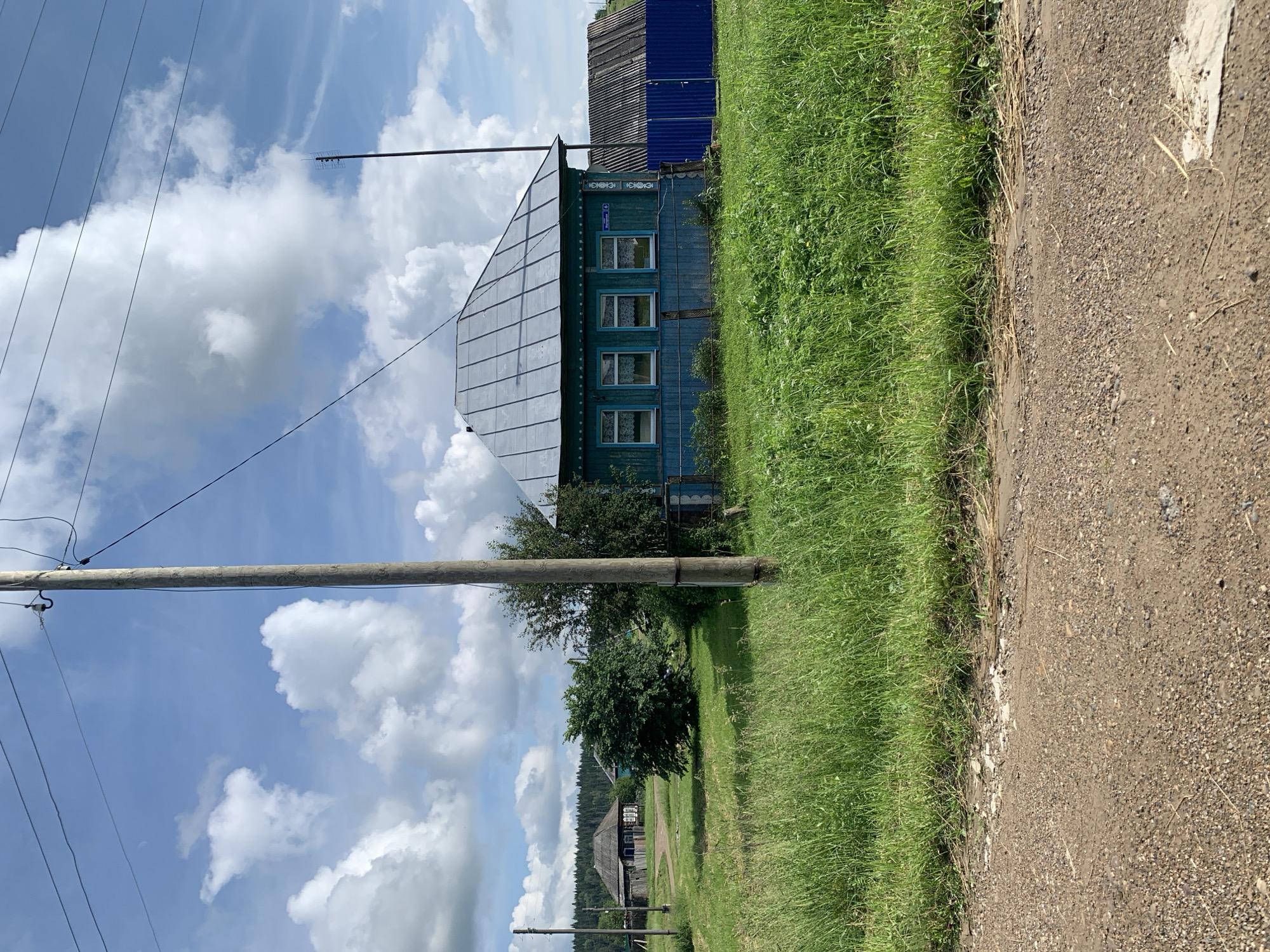 The image captures a rural scene in Barsai, Russia, on a sunny day with scattered clouds. In the foreground, a dirt road leads into a grassy field. To the right, a traditional blue wooden house with a gray metal roof stands prominently. The house features three visible windows with white lace curtains. A blue fence encloses part of the property. Further back, a wooden utility pole with power lines crosses the frame. In the background, other rural houses and dense green trees are visible, suggesting a small village or hamlet. The overall atmosphere is peaceful and idyllic.