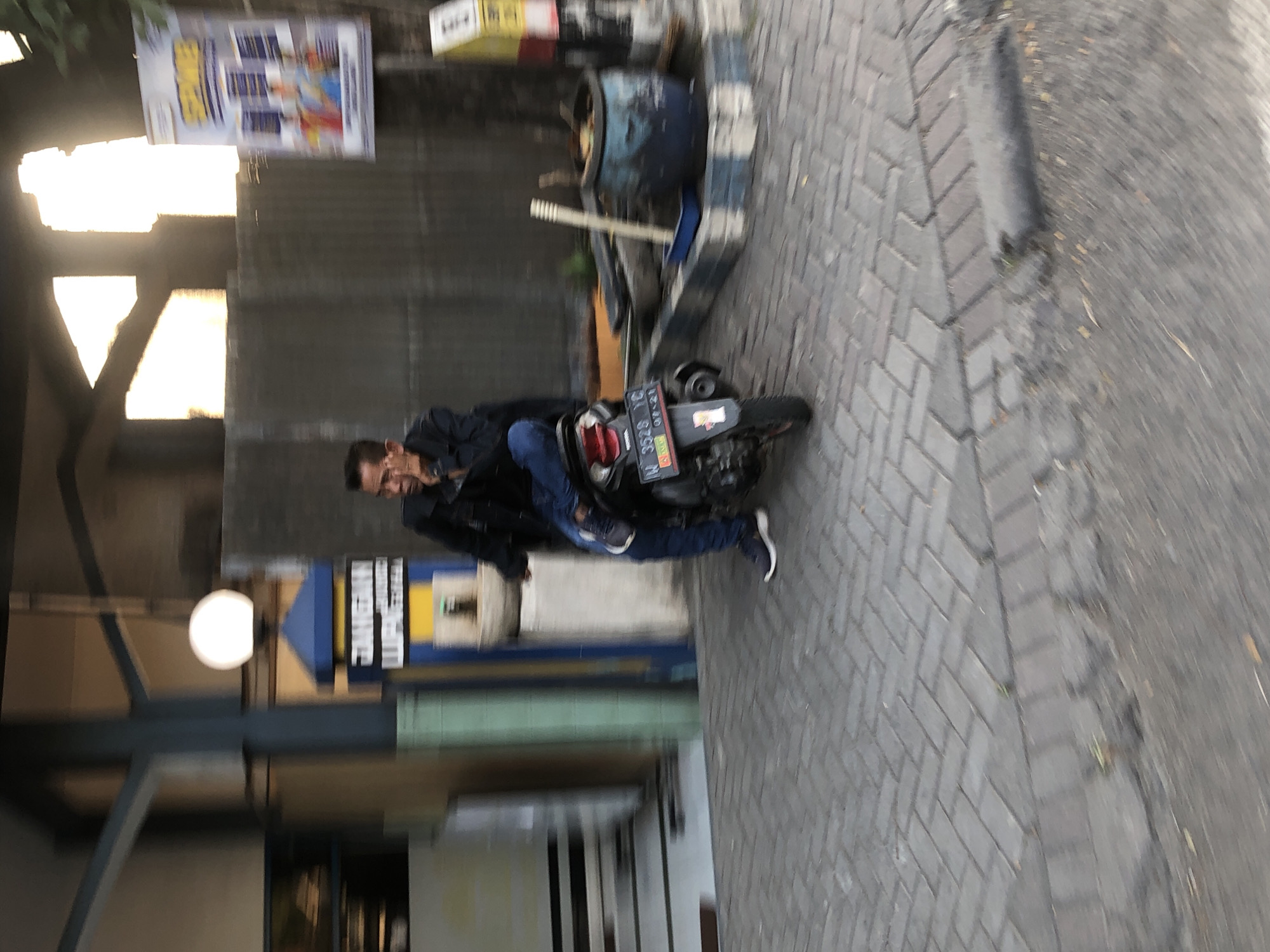 A man is sitting on a motorcycle in Ngaban, Indonesia. He is wearing a dark jacket and blue jeans. The motorcycle is black and has a license plate that reads "W 3528 YC". In the background, there is a building with a blue and yellow sign. There is also a large blue pot and a broom. The ground is paved with interlocking bricks. The image is blurry and appears to have been taken while moving.