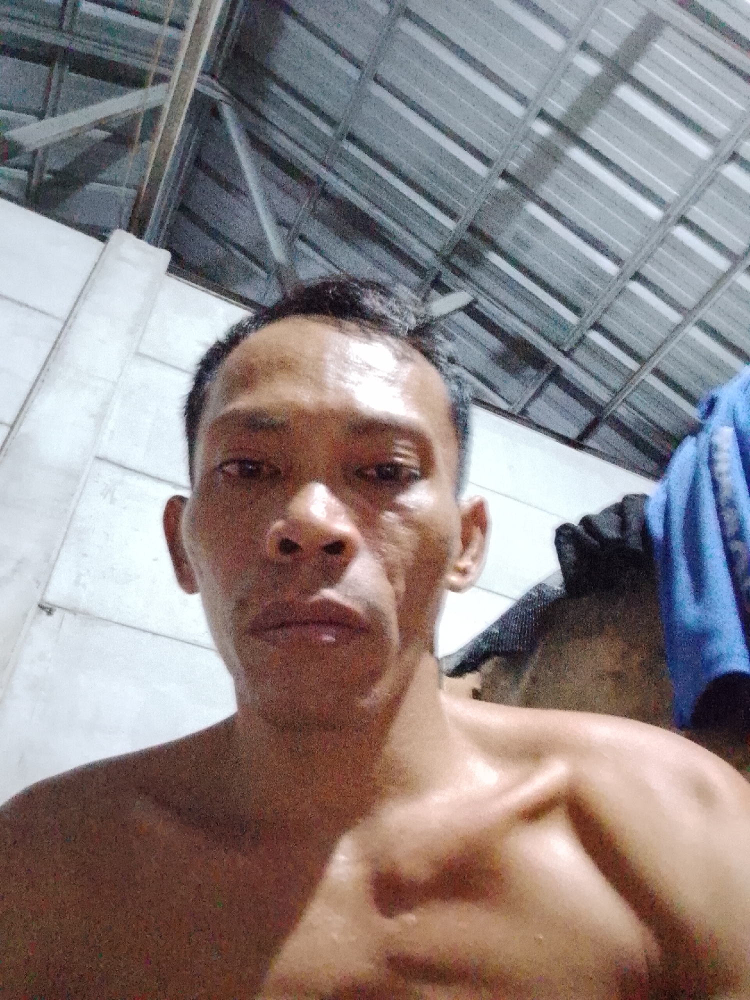 A shirtless man is shown in a close-up selfie. He has short, dark hair and is looking directly at the camera. He is fair-skinned. The background appears to be an interior space with a ceiling made of corrugated metal and a white wall in the background. A blue cloth is partially visible in the background. The photo appears to be taken indoors in Bukit Ketok, Indonesia.