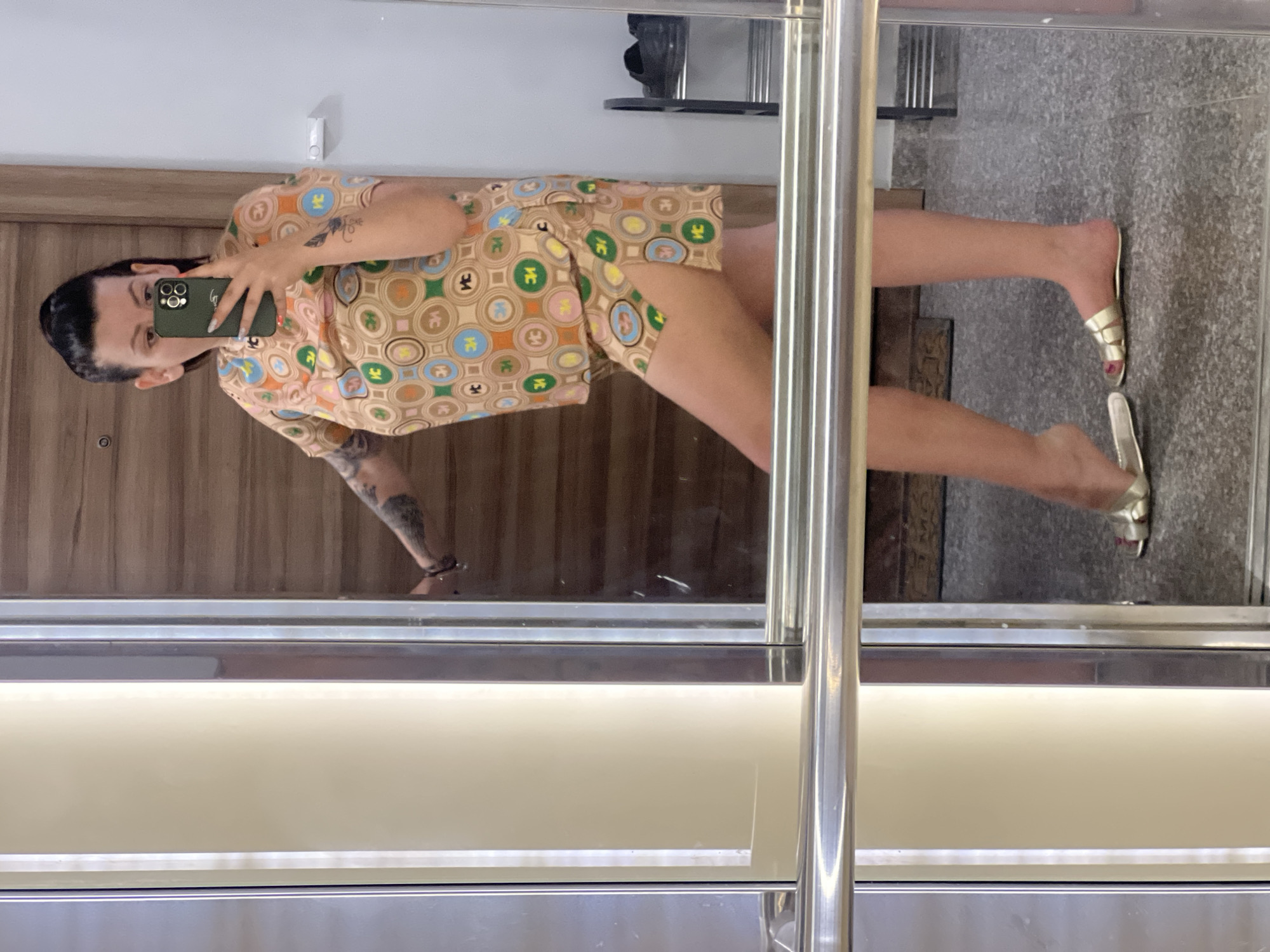 A woman is taking a mirror selfie in an elevator. She is wearing a light brown short-sleeved pajama set with a colorful circular pattern. The pajamas have a collar and short shorts with slits on the sides. She has tattoos on her arms. She is holding a green smartphone in her right hand, facing the mirror. She is wearing gold slide sandals. The elevator has stainless steel walls and a mirrored surface that reflects the woman and the surrounding area. In the reflection, a wooden door is visible behind her, along with a grey floor. A black shoe rack with two pairs of black shoes is also visible in the reflection. The overall setting appears to be a modern residential building, potentially in the city of Plovdiv, Bulgaria, given the context of the request.