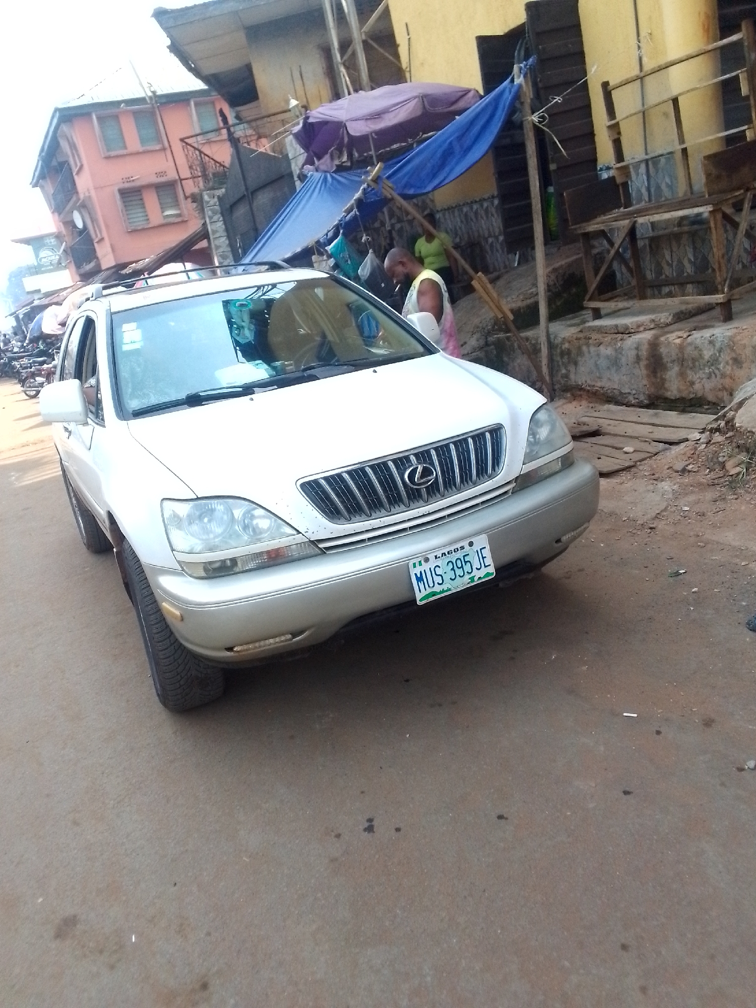 A white Lexus SUV is parked on a paved street in Orlu, Nigeria. The license plate reads "MUS 395 JE" and identifies the state as Lagos. The car is angled towards the viewer. The background shows a mix of residential and commercial buildings. There are people under blue tarps attached to storefronts, suggesting a market area. The overall environment appears urban, with signs of wear and tear. Some motorcycles are seen parked nearby.