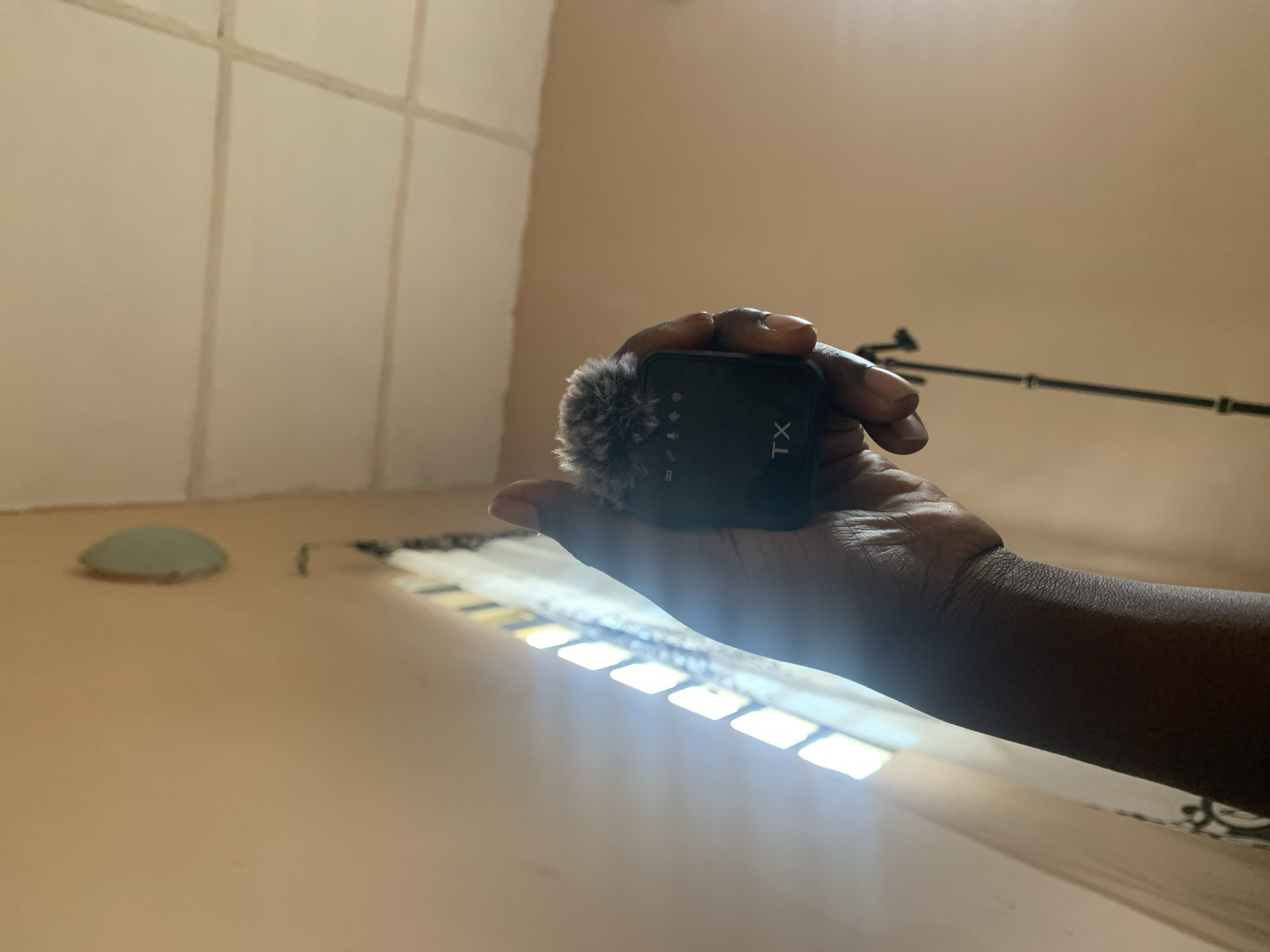 An over-the-shoulder, low-angle shot captures a dark-skinned hand holding a black wireless microphone with a furry windscreen. The microphone has the letters "TX" in white on its body. In the background, a light-colored wall and ceiling are visible. A window with horizontal blinds is positioned to the left, allowing sunlight to stream in, creating a lens flare effect. A black tripod is partially visible below the microphone. The overall lighting suggests an indoor environment during the day. The setting is not identifiable as being in a specific city.