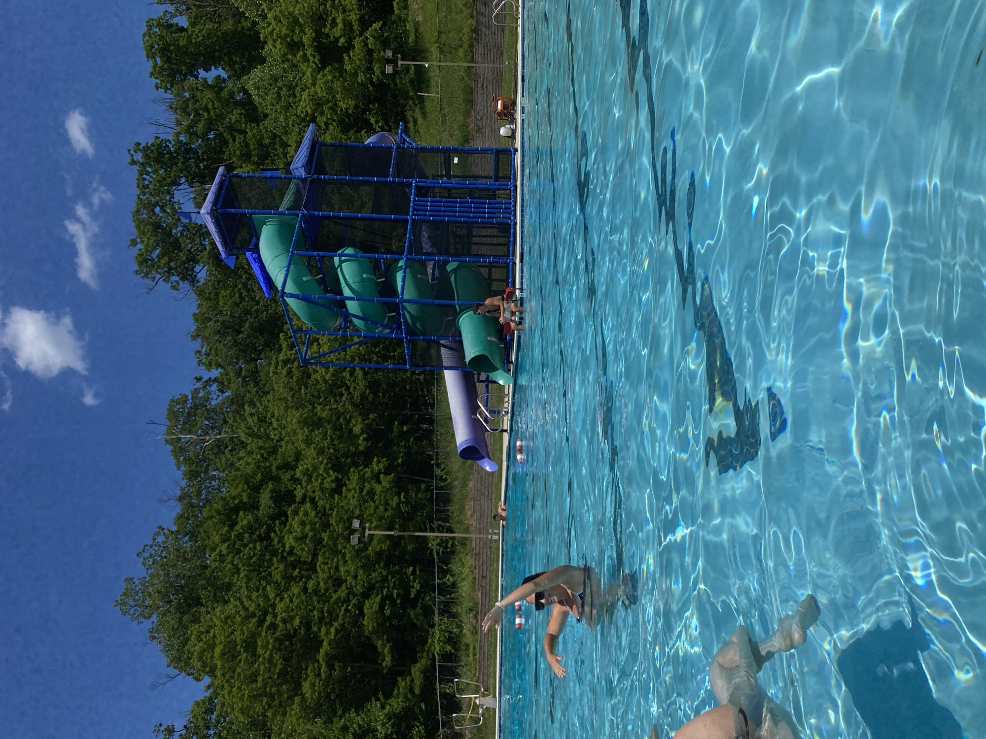 In Washington Township, United States of America, a bright, sunny day is captured at an outdoor swimming pool. The pool's clear blue water is filled with shimmering light reflections. In the foreground, a woman in a dark swimsuit and sunglasses is in the water, with her left arm raised. To her right, a portion of another person's legs are visible above the water.
In the background, a large, multi-level water slide structure stands prominently. This structure features a blue frame with several green tube slides and one purple tube slide. People are visible near the slide, with one person sitting on the edge of the pool near the slide's exit. The area surrounding the pool is lined with concrete, and beyond that, a dense green forest stretches towards a bright blue sky dotted with white clouds. The scene conveys a sense of summer recreation and enjoyment.