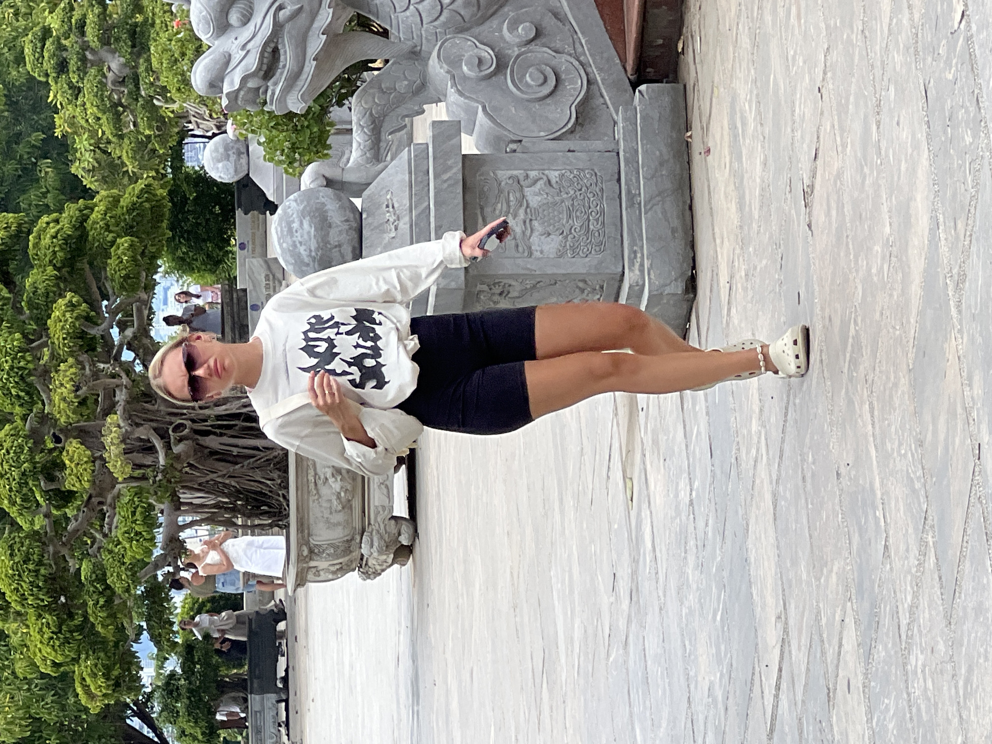 The image shows a young woman walking towards the camera in an outdoor paved area. She is wearing a white long-sleeved shirt with a black graphic print, black shorts, white Crocs, and sunglasses. She is holding a mobile phone in her right hand and a white purse over her left shoulder. In the background, there is a large tree, stone sculptures including a dragon head, and other people walking. The location is identified as the Marble Mountains in Đà Nẵng, Vietnam, an area known for its Buddhist temples and pagodas carved into the limestone hills.
