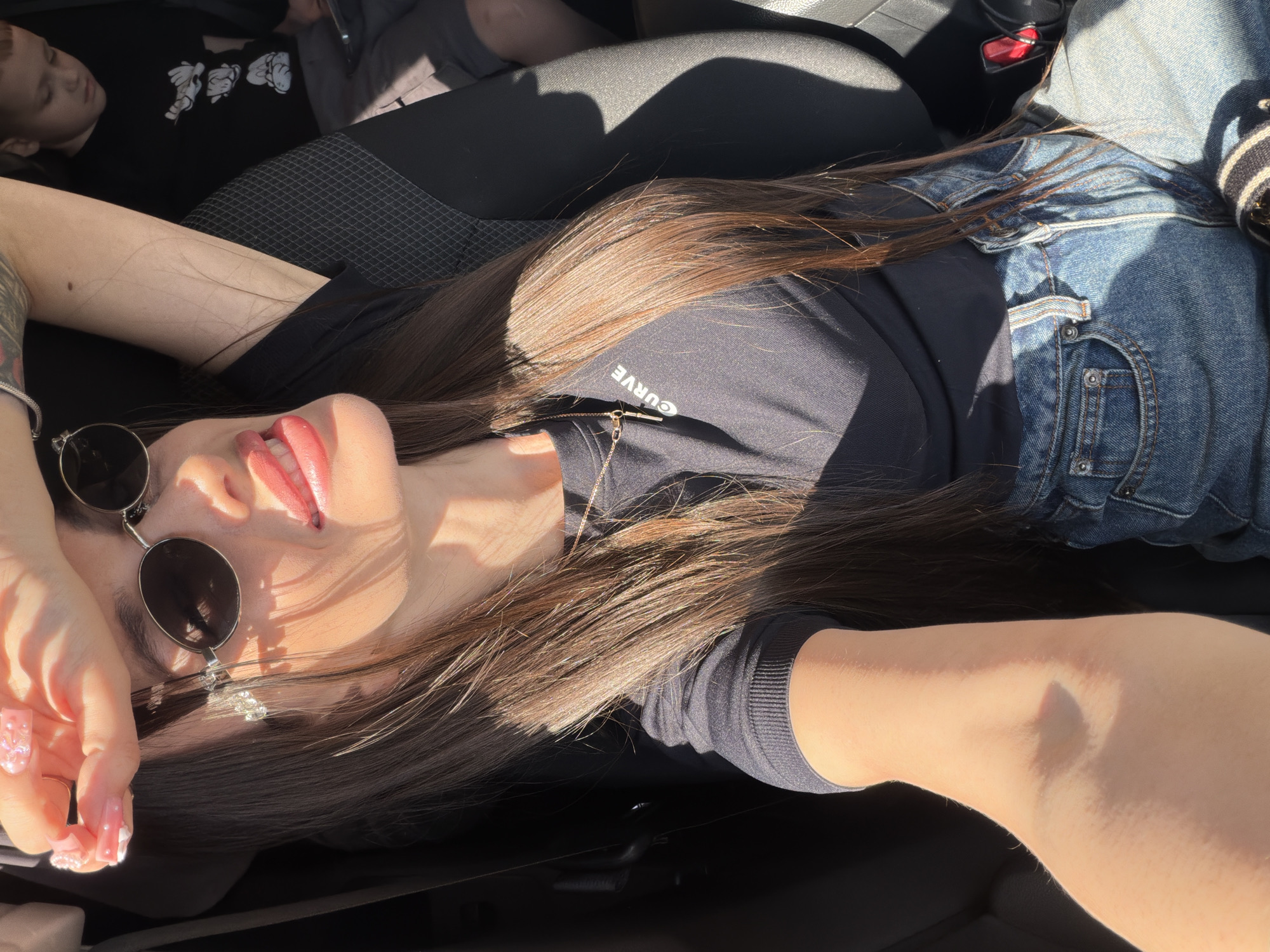 The image captures a woman and a child inside a car on a bright, sunny day in Izhevsk, Russia. The primary subject is a woman, positioned lying across the car seats with her head tilted back. She wears a black long-sleeved top, featuring the word "CURVE" printed upside down on the chest, and blue denim jeans. Round sunglasses are perched on her face, and her long, dark brown hair is spread out. Her hands are adorned with clear and pink ornate acrylic nails, with one hand resting near her face. A delicate gold chain necklace is visible around her neck.
In the background, a child lies on another car seat, wearing a black t-shirt with white cartoon characters. The child appears to be sleeping or resting.
Strong, direct sunlight streams into the car, creating prominent highlights on the subjects' skin, hair, and clothing, and casting distinct shadows. The car interior features dark-colored seats with a textured fabric pattern. A red seatbelt buckle is partially visible. The scene evokes a sense of relaxation or a casual, sun-drenched moment, possibly during a car ride or a stop.
