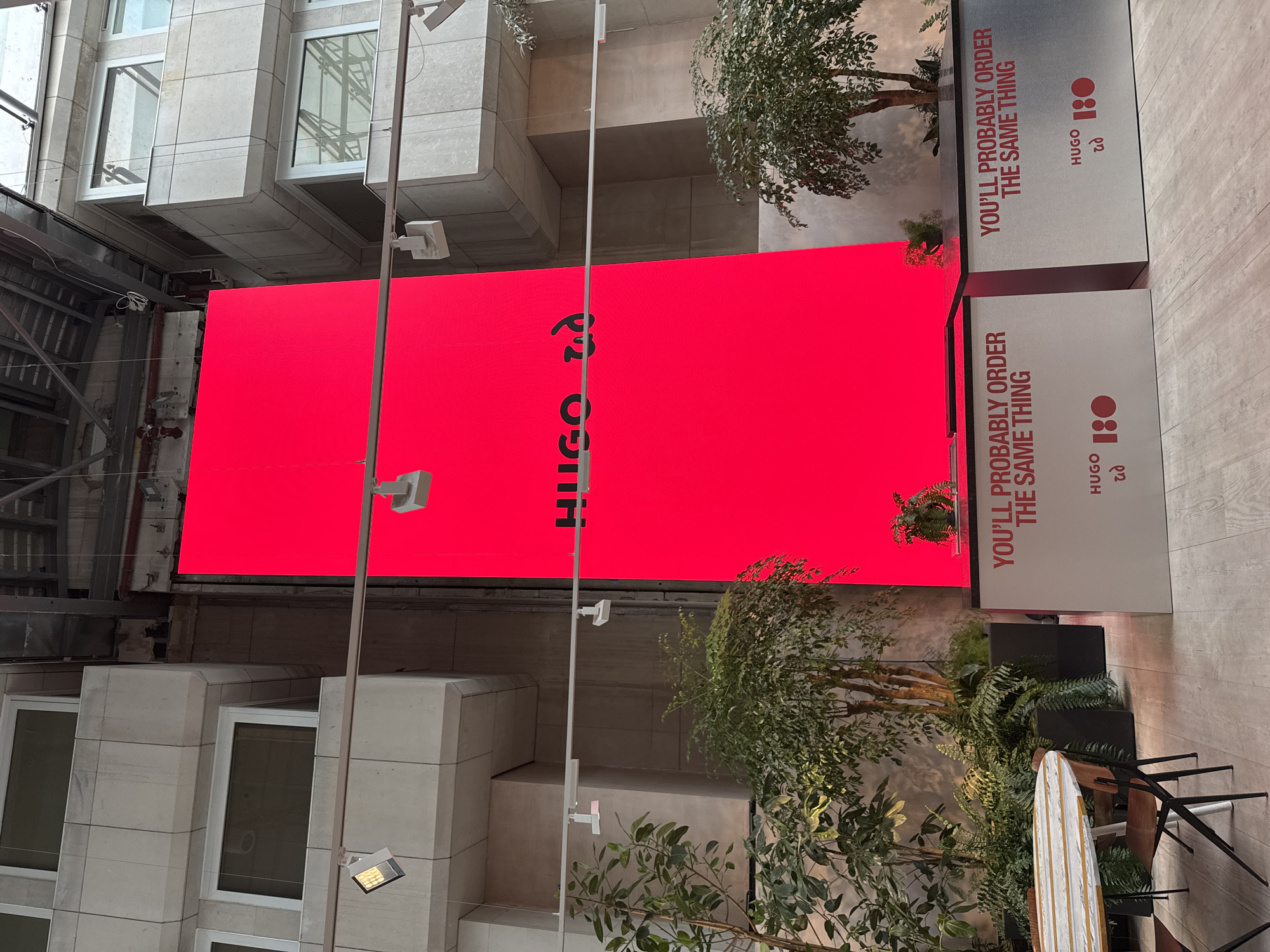 The image shows a Hugo Boss advertising display in a city setting, likely London, United Kingdom, based on the style of architecture. The display includes a large red screen with the Hugo Boss logo vertically oriented and text that reads "YOU'LL PROBABLY ORDER THE SAME THING," also a logo with "HUGO" and "1:0" on a white background. The scene includes modern architecture with block-shaped buildings and visible windows. There are street lamps and foliage incorporated into the design, with trees or plants in the foreground, suggesting an urban environment with attempts at greenery. There are also a table and chairs suggesting seating area for customers.