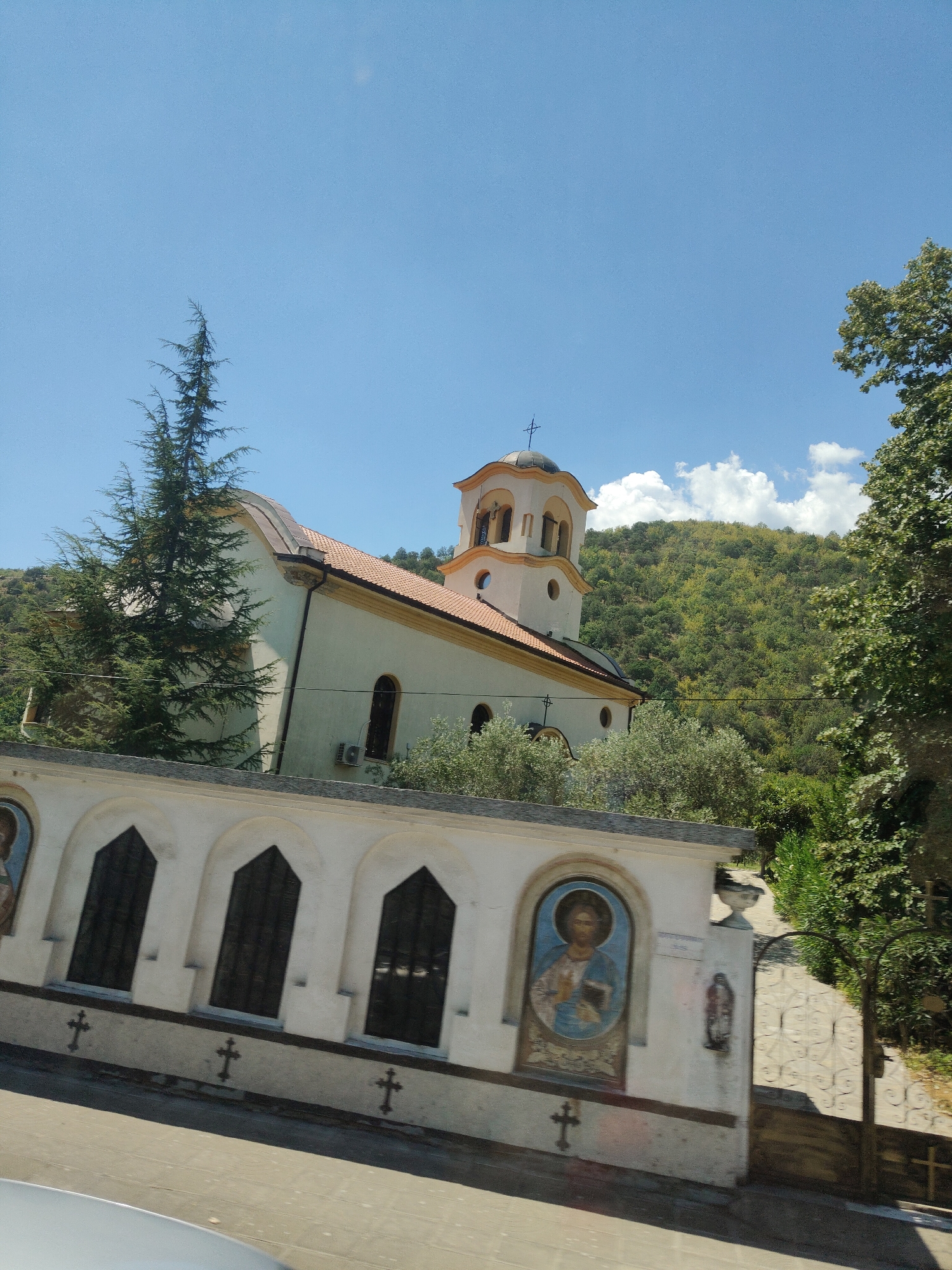 In Kresna, Bulgaria, an Eastern Orthodox church is nestled against a lush, green mountainside under a clear blue sky. The church features a light yellow facade, a terracotta-tiled roof, and a prominent bell tower topped with a cross. A decorative white wall in the foreground displays several arched windows and religious iconography, including images of Jesus Christ. Green trees and foliage surround the church, contributing to the natural beauty of the setting. The scene is captured from a slightly angled perspective, suggesting the photograph was taken from a moving vehicle.