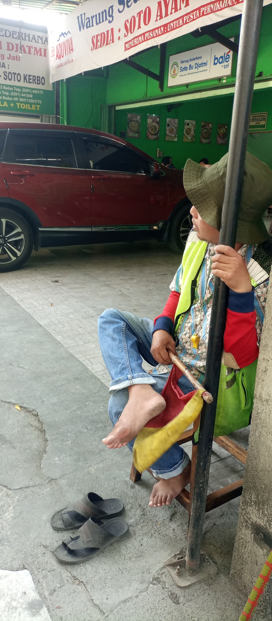 In Jatirejo, Indonesia, a person sits on a wooden chair near a street. They are wearing a hat, a colorful shirt, jeans, and a neon yellow vest. Their left foot is bare and appears dirty. In their hand, they hold a red and yellow flag attached to a wooden stick. Beside them, a pair of dark-colored sandals rests on the ground. Behind them, a red car is parked. Various signs are visible in the background, one of which reads "Warung Sederhana Soto Bu Djatmi." The environment suggests a roadside eatery or a business establishment.