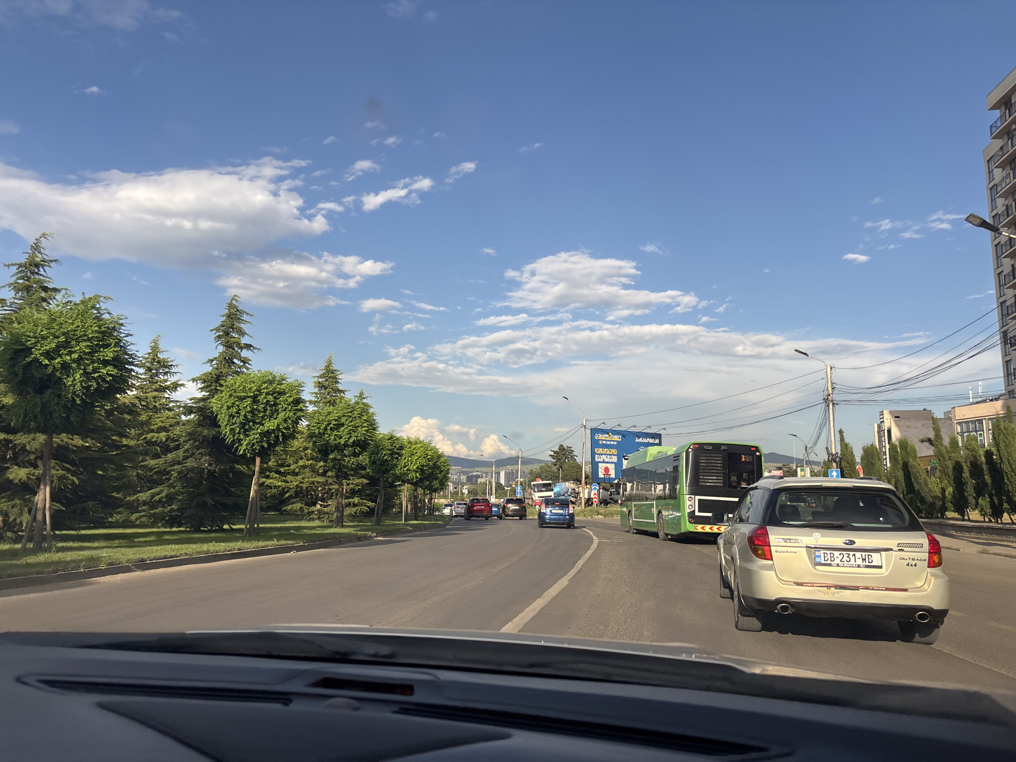 This media file depicts a street scene in Tbilisi, Georgia, likely during the daytime under a partly cloudy sky. The perspective is from inside a vehicle, looking forward through the windshield.
The primary subjects are vehicles on a multi-lane road. In the foreground, the dashboard of the vehicle the photo is taken from is visible. Further down the road, there are several cars, including a silver Subaru Outback with a license plate reading "BB-231-WB". A large green bus is also prominent, positioned to the right of the Subaru. To the left of the road, there's a grassy area with well-maintained trees, some of which are conical or spherical in shape, suggesting a park or landscaped area. In the background, there are buildings and more trees, with hills or mountains visible in the distance.
The activity taking place is traffic on a road. The vehicles appear to be moving, suggesting an everyday commute or travel. The overall mood of the scene is calm and ordinary, with the bright sky and lush greenery contributing to a pleasant atmosphere.
Notable details include the clear blue sky with scattered white clouds, characteristic of good weather. The presence of traffic lights and streetlights indicates an urban or suburban environment. A large billboard with text in Georgian is visible in the background, offering a specific location cue. The text on the billboard appears to be advertising, with the word "ინტერნეტი" (internet) and a phone number visible. The Subaru Outback also has text on its rear, reading "SUBARU OUTBACK 4X4". The time of day appears to be late afternoon or early evening, judging by the quality of light and the shadows.
