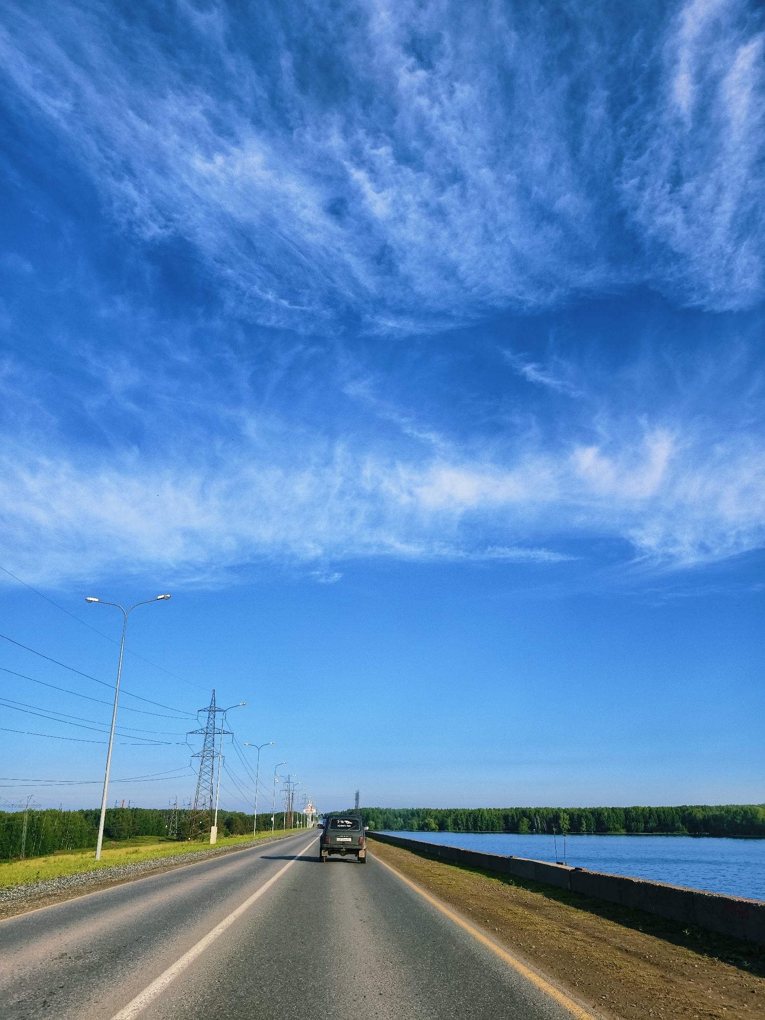 The image captures a scenic view from a moving vehicle on a clear, sunny day in Chaikovsky, Russia. A vast, bright blue sky dominates the upper portion, adorned with delicate, wispy white clouds scattered across its expanse.
In the mid-ground, an asphalt road stretches into the distance, with a dark-colored car, possibly an SUV, driving ahead and away from the viewer. The car has a sticker on its rear window with partially visible text, possibly "ПЕГАС-НО" (PEGAS-NO). The road features a solid white line marking the shoulder and a double solid yellow line down the center, indicating two-way traffic.
To the left of the road, a strip of green grass borders the asphalt, followed by several utility poles with power lines and a large metal power pylon. Beyond these structures, a dense line of lush green trees forms a forest, stretching into the horizon.
To the right of the road, a low concrete barrier separates the road from a wide expanse of calm, blue water, likely a river or lake (consistent with Chaikovsky's location on the Kama River). Another dense line of green trees can be seen on the opposite bank of the water body, completing the natural backdrop. The overall scene suggests a peaceful journey through a rural or semi-rural landscape.